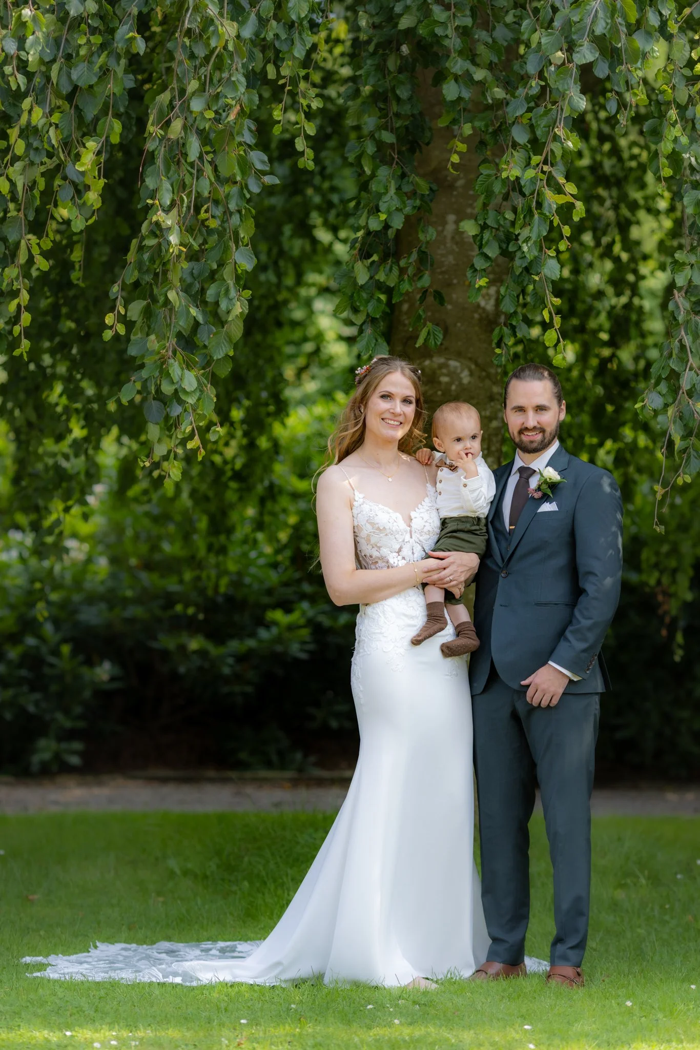 Et brudepar med en baby poserer foran et træ i en grøn have.