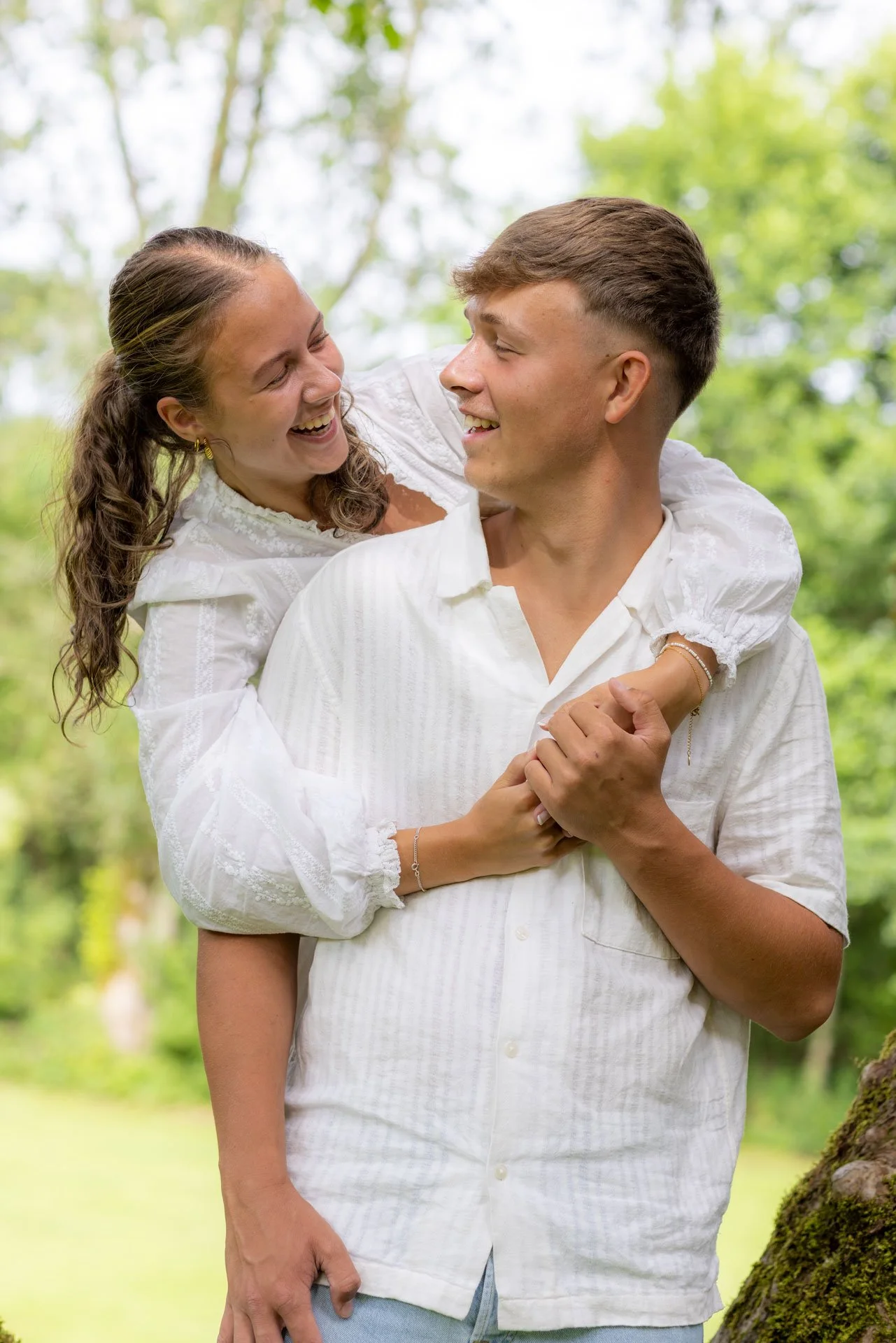 Et smilende par, hvor kvinden sidder på mandens ryg, begge er iført hvide skjorter, i en grøn skov.