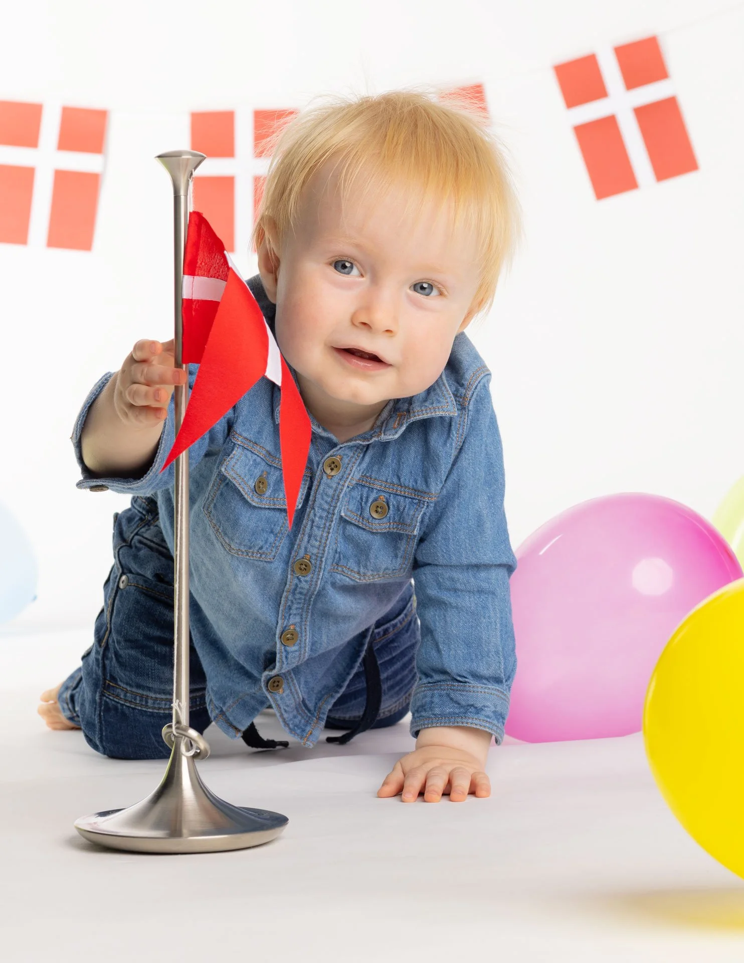 En lille dreng med blond hår iført denimjakke og bukser, der leger på gulvet med balloner og en dansk flag-stang. Der er danske flag i baggrunden.