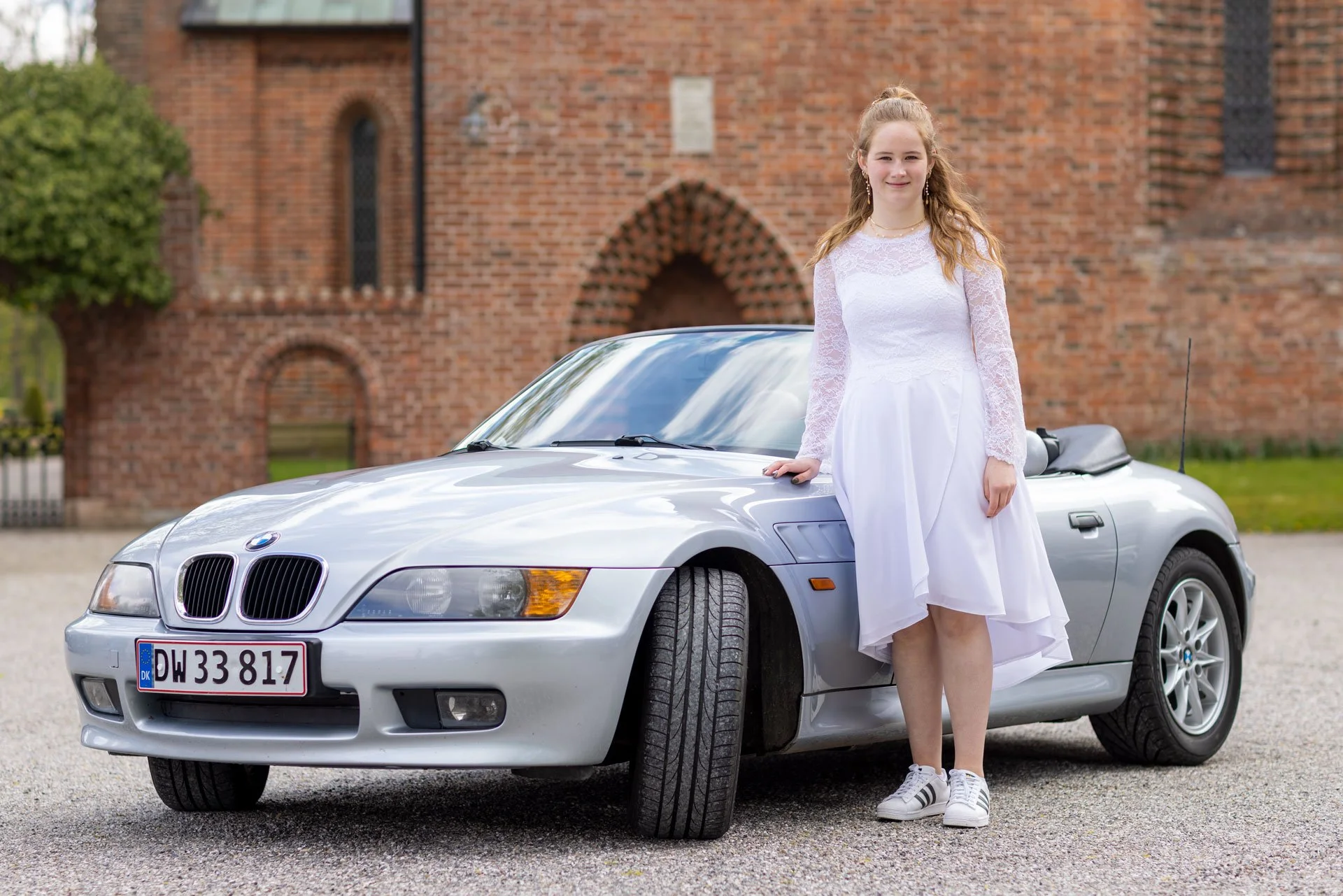En ung kvinde i en hvid kjole og sneakers står ved siden af en sølv BMW convertible foran en rød murstensbygning.