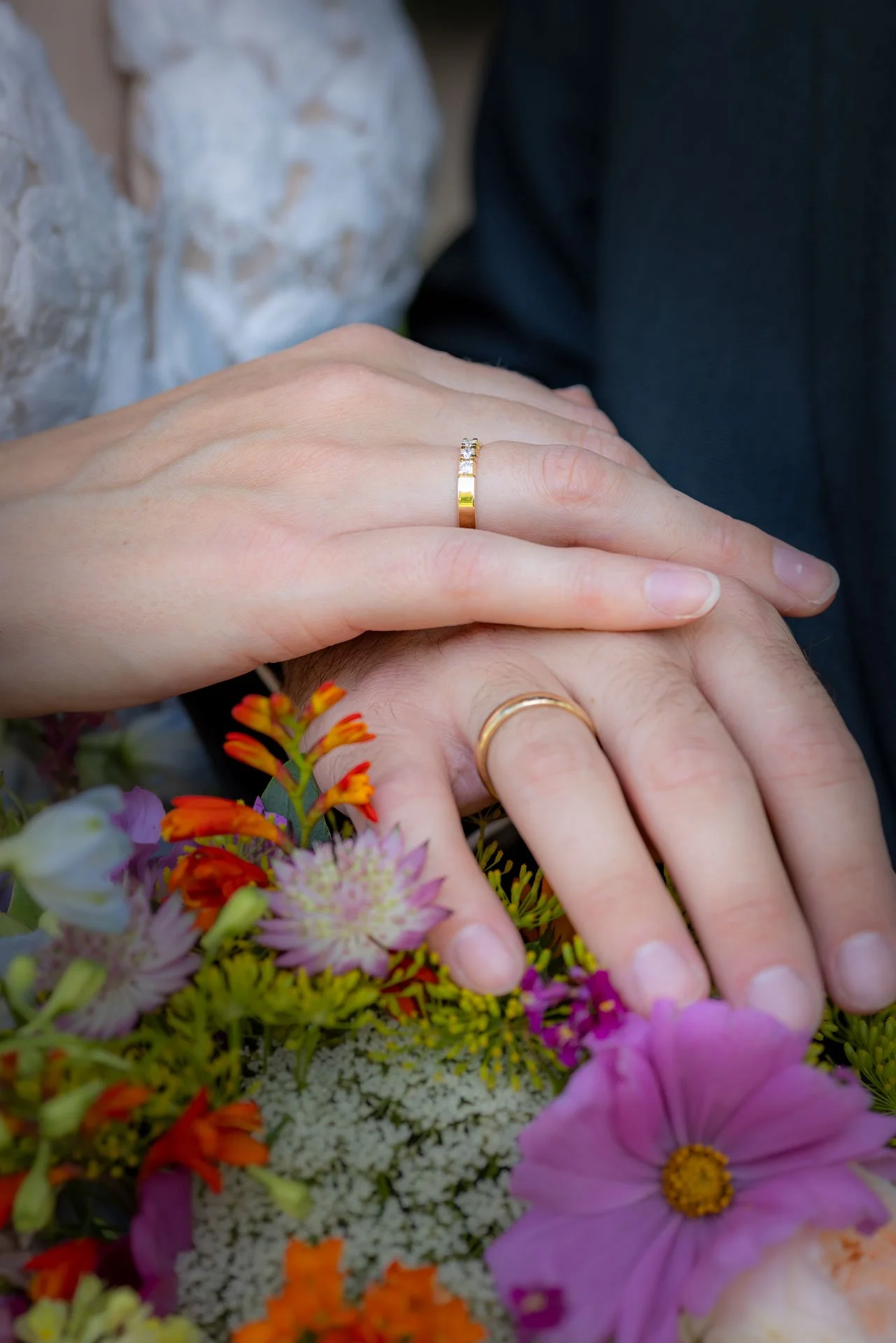 Billedet viser to hænder, der viser deres guld- og diamantbrydende vielsesringe, placeret ovenpå en bunke af farverige blomster. En person har en diamantbesat ring, mens den anden har en enkel guldring.