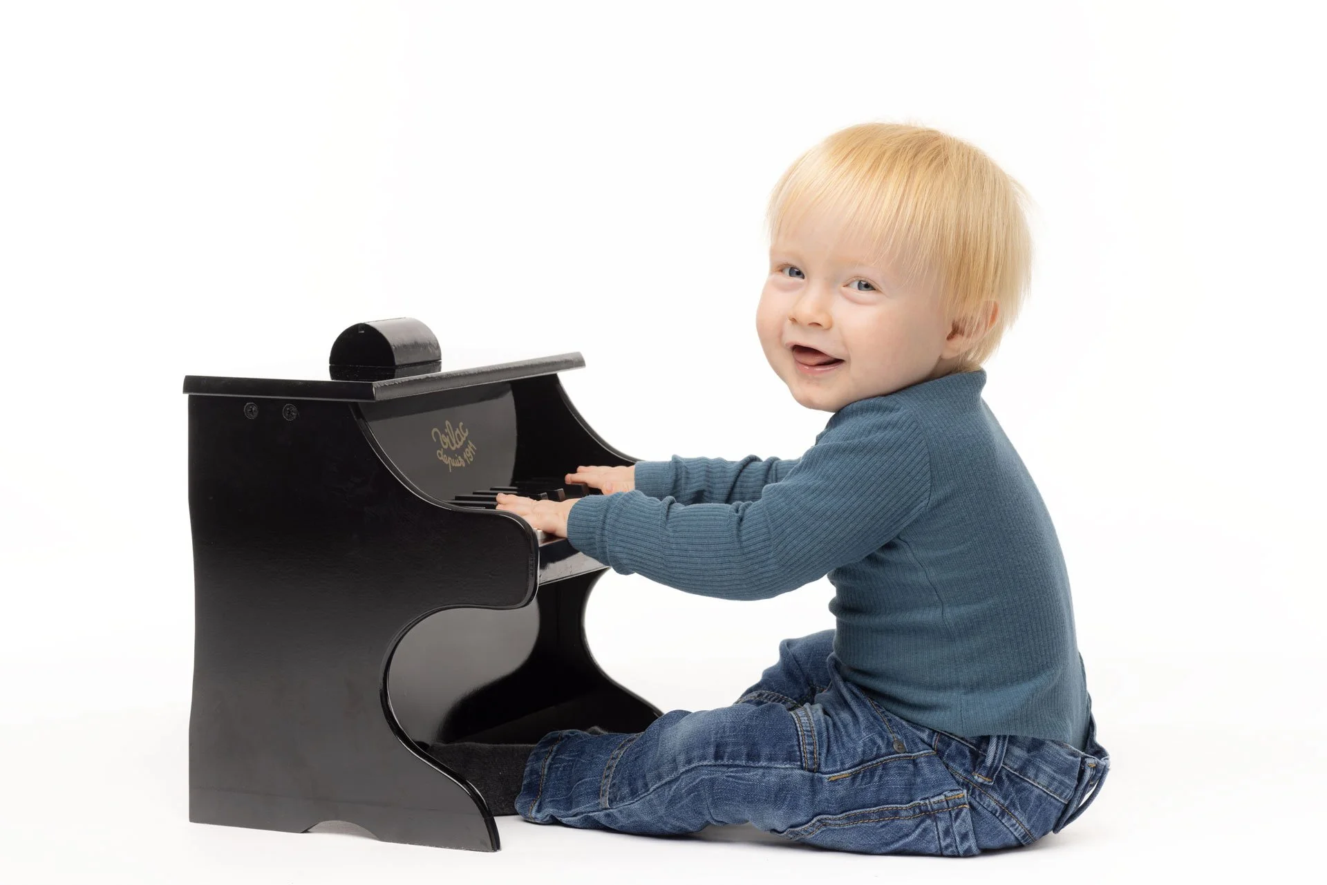En glad blond dreng sidder på gulvet og spiller på en lille sort legetøjspiano mod en hvid baggrund.