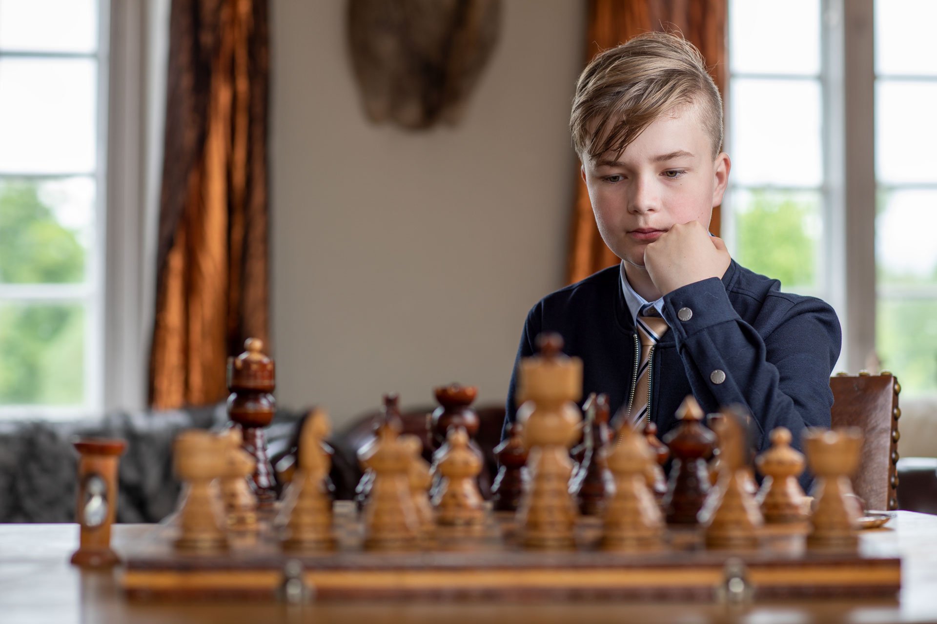 Ung dreng spiller skak ved et bord i et rummeligt værelse med store vinduer.