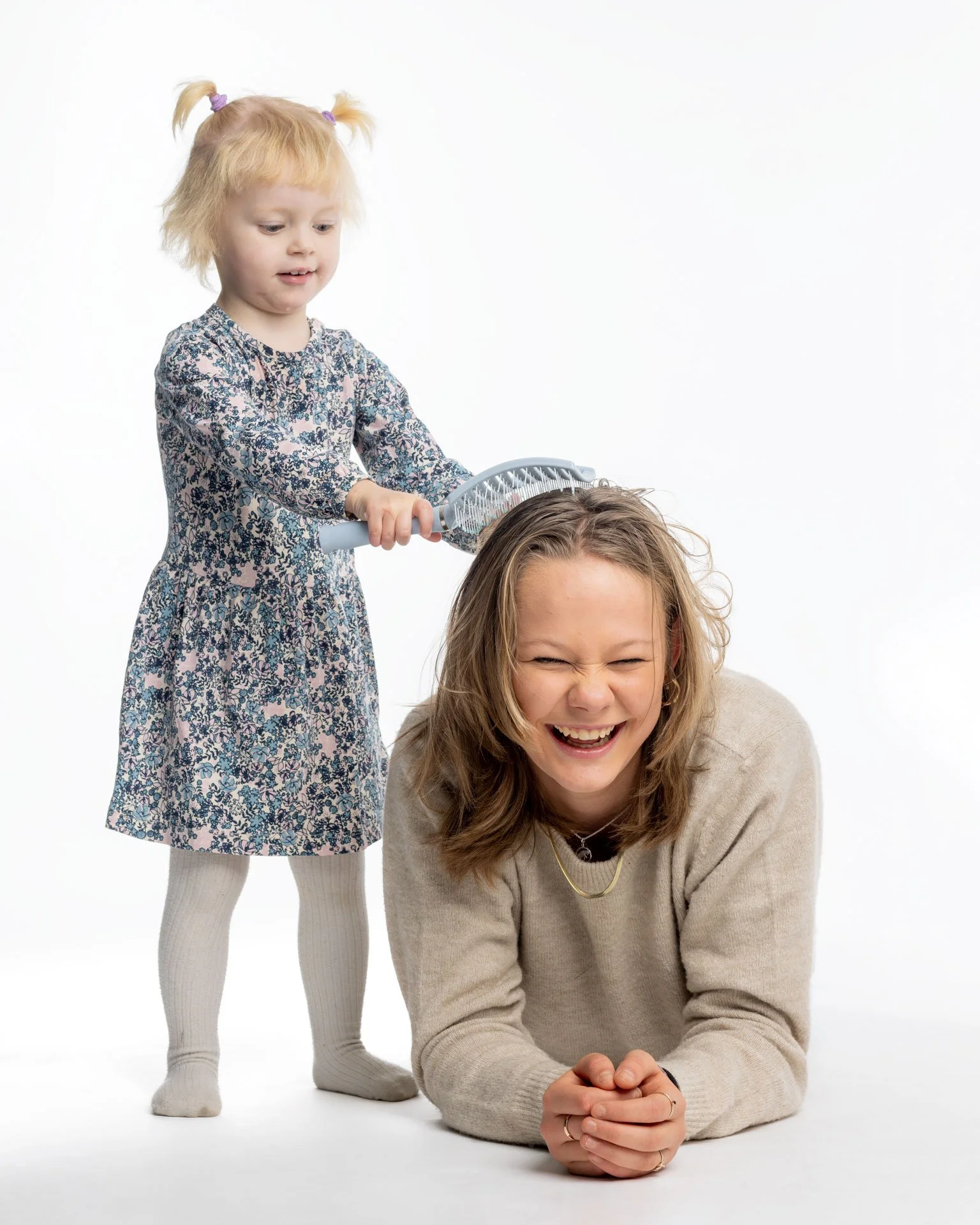 En lille pige med blonderhår, der leger, at hun børster en voksen kvindes hår, der ligger på gulvet, og begge smiler og griner.