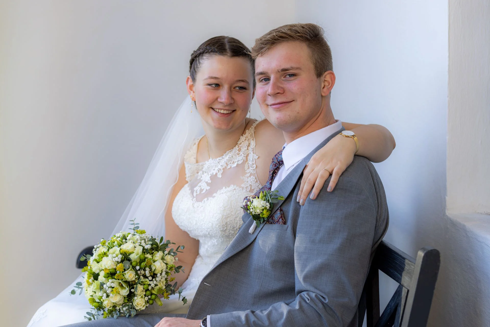 Et brudepar sidder på en bænk, med bruden i en hvid brudekjole og brudgommen i en grå habit, begge smilende.