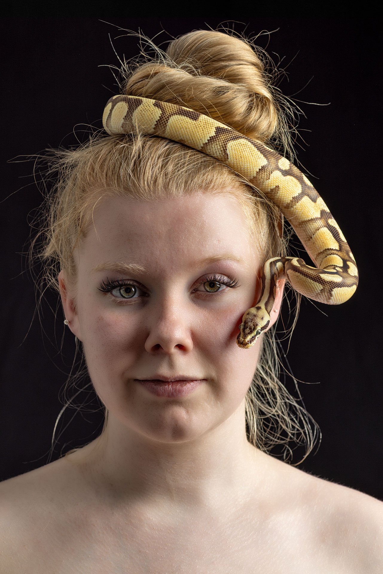 Portræt af en ung kvinde med blond hår i en oppustet hårbølle, med en stopklædt slange snorende sig over hovedet og skulderen, mod en sort baggrund.