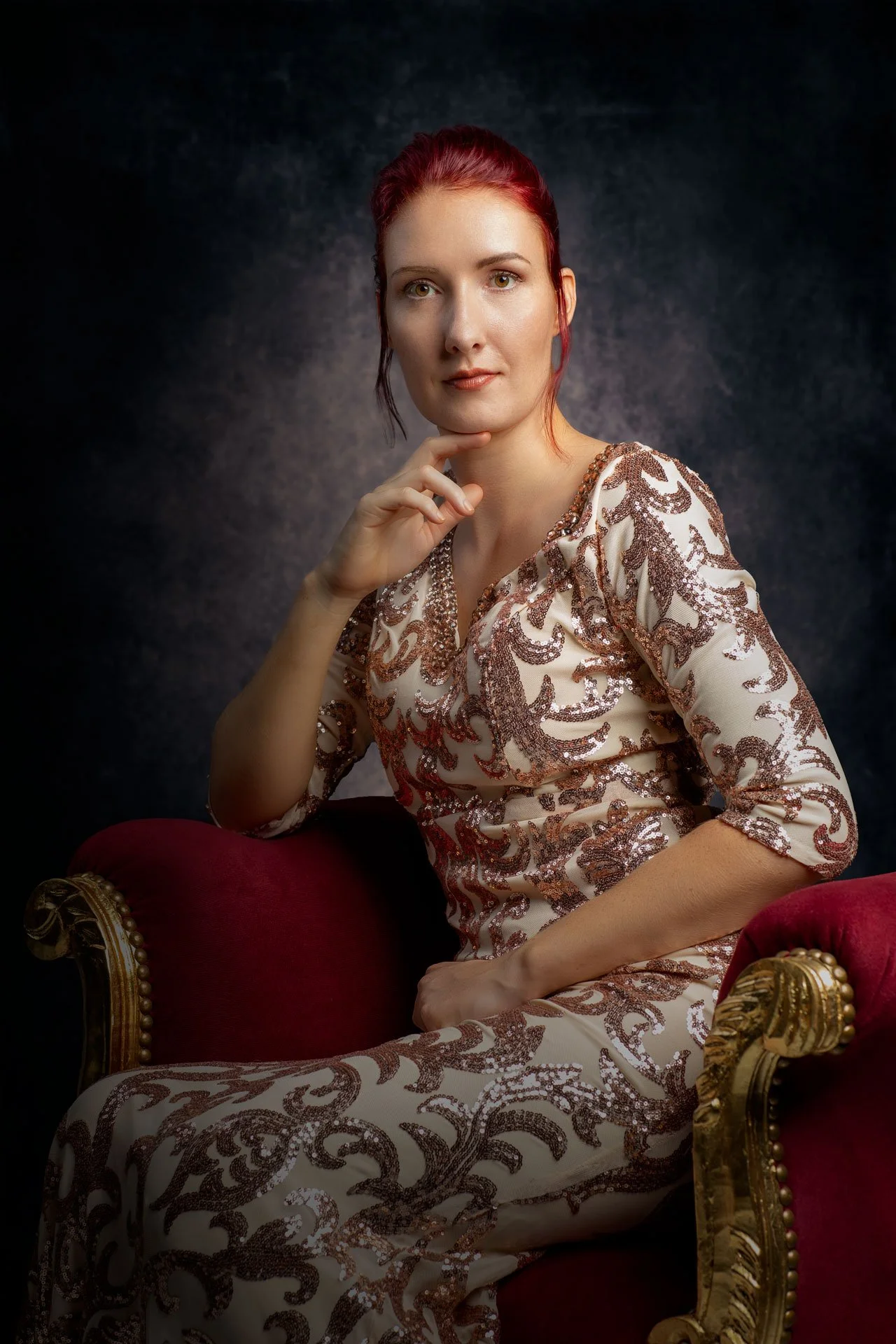 En kvinde med rødt hår sidder i en rød, gylden stol iført en beige kjole med broderede mønstre.