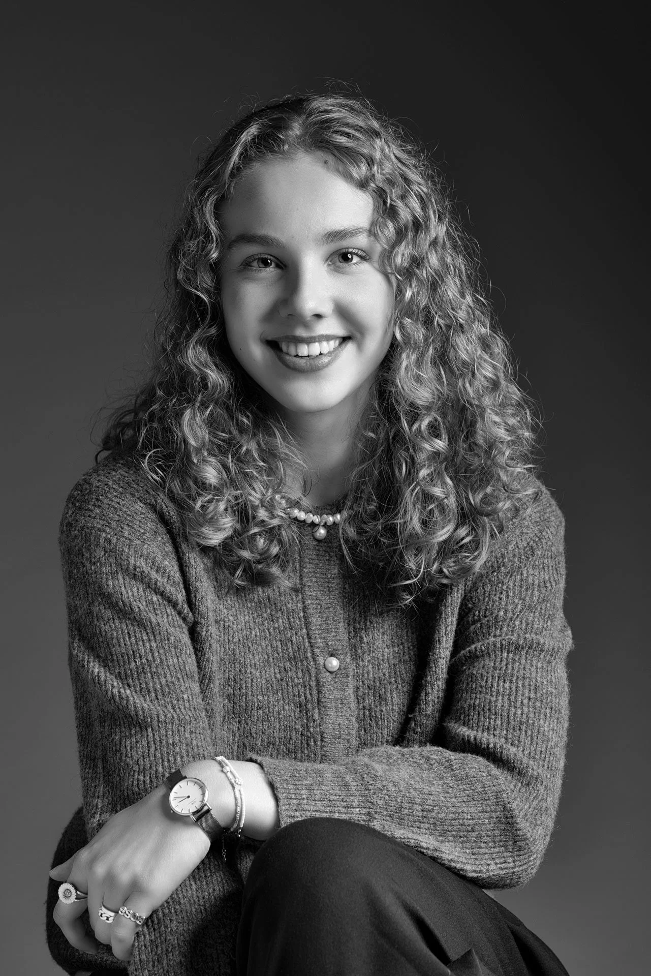 Sort-hvid portrætfotografi af en smilende kvinde med krøllet hår, iført strikket cardigan, smykker og ur, siddende med armene krydsede.