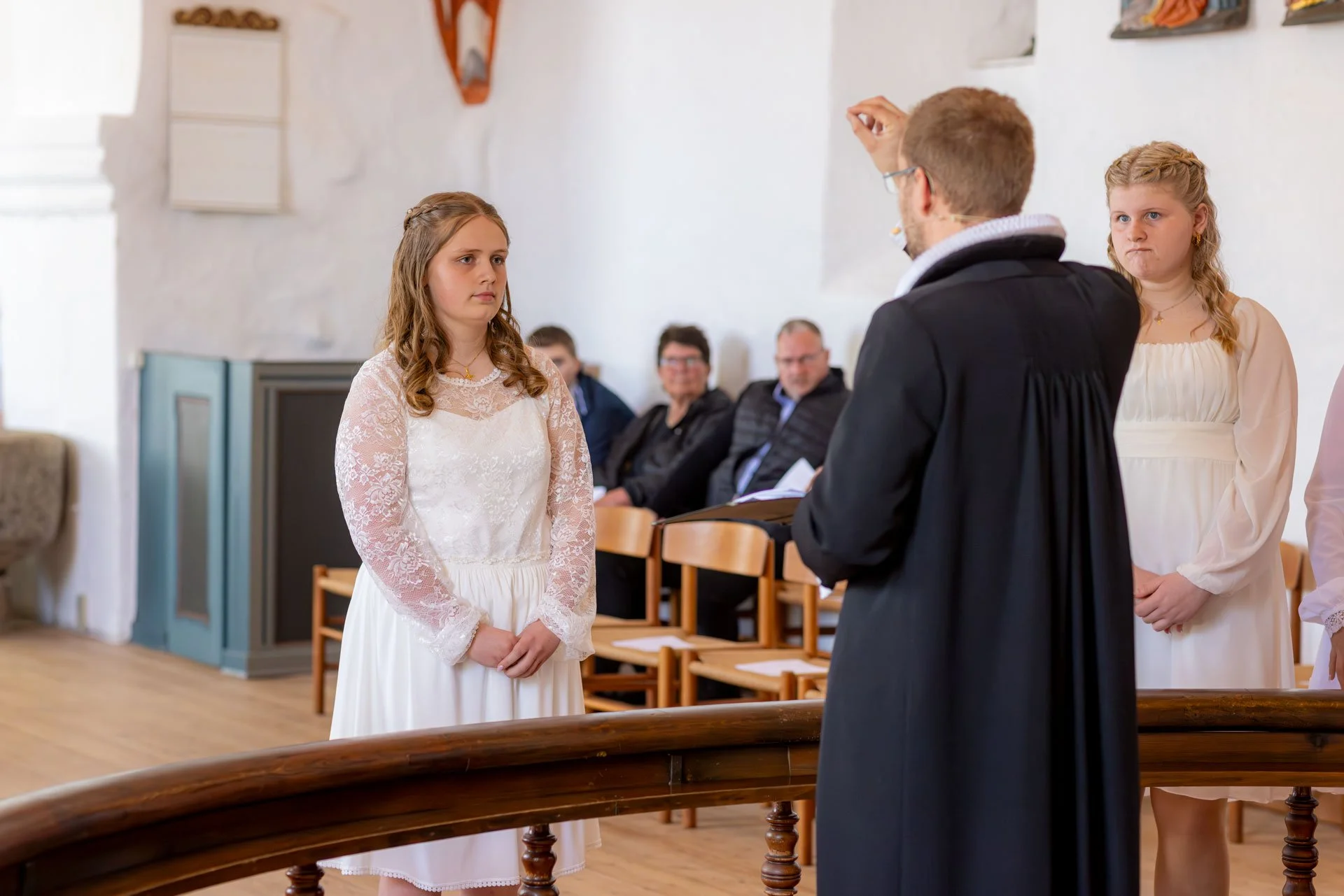 En person iført en sort præstekjole står foran to unge piger i hvide kjoler, der deltager i en ceremoni i kirke med tilhørere i baggrunden.