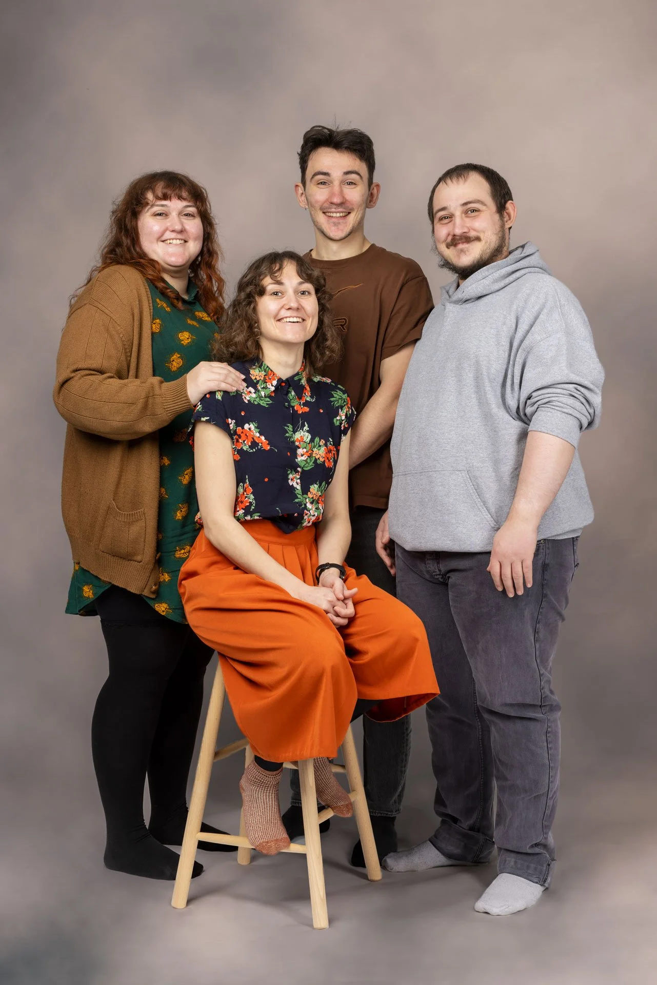 Fem personer poserer for et foto mod en neutral baggrund, hvoraf en sidder på en stol. De smiler og er klædt i afslappet tøj.