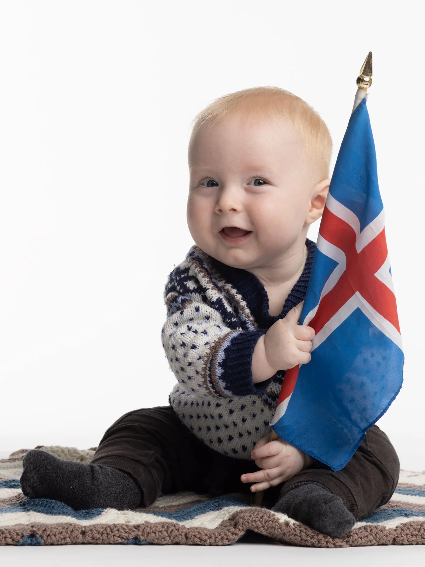 En lille dreng med blond hår, der sidder på en vævet tæppe og holder et Islandsk flag.