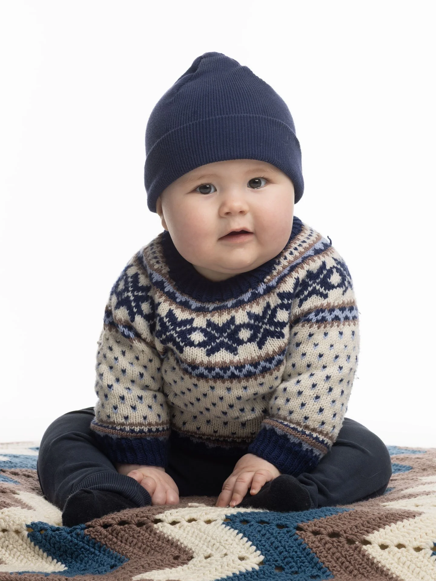 Sødt baby sidder på gulvtæppe, iført strikket sweater med nordisk mønster, blå hue og sorte bukser.