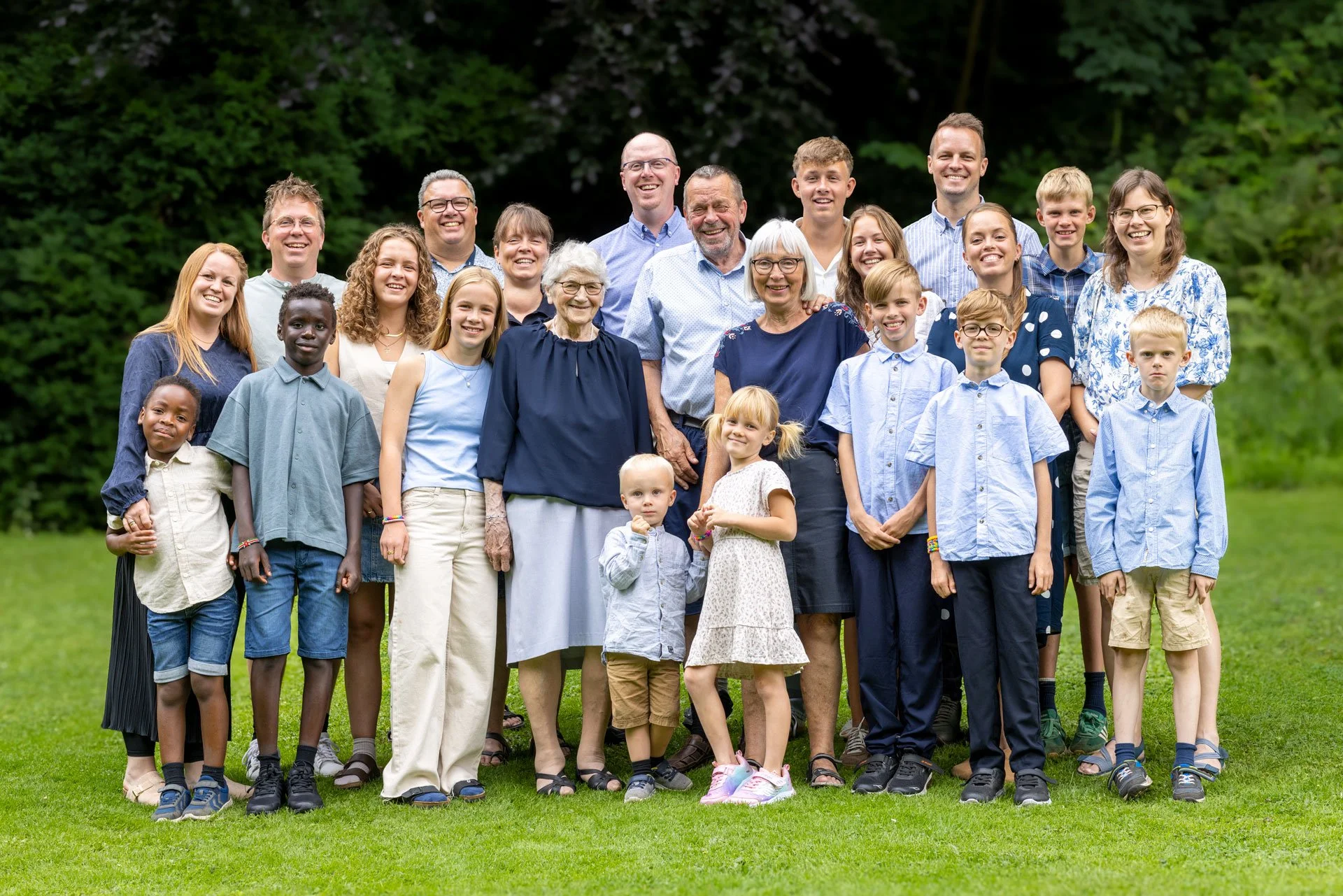 En gruppe mennesker, inklusive børn og ældre, poserer udendørs på en grøn græsplæne med træer i baggrunden.
