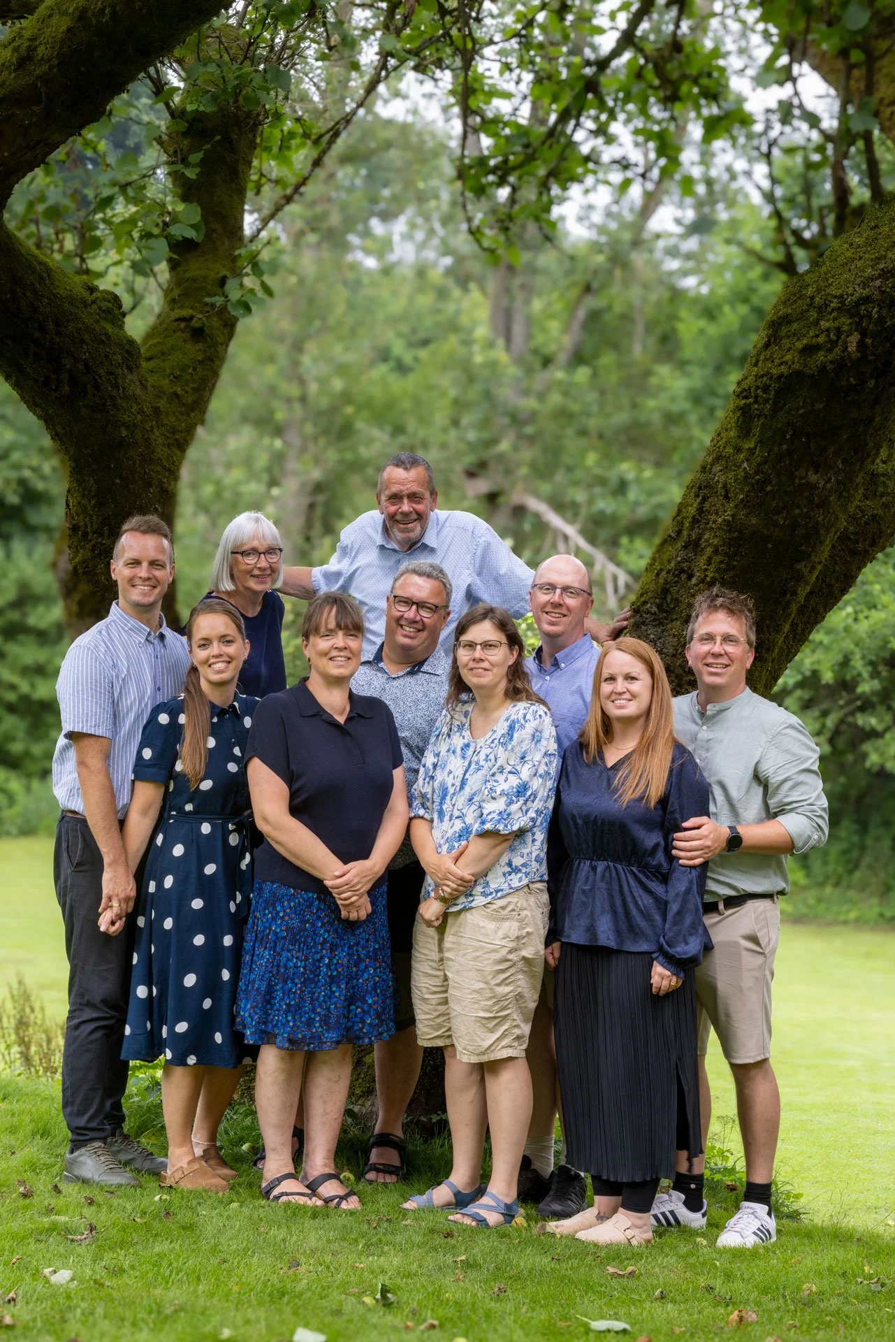En gruppe mennesker står sammen under et træ i en grøn have. De smiler og poserer for kameraet.