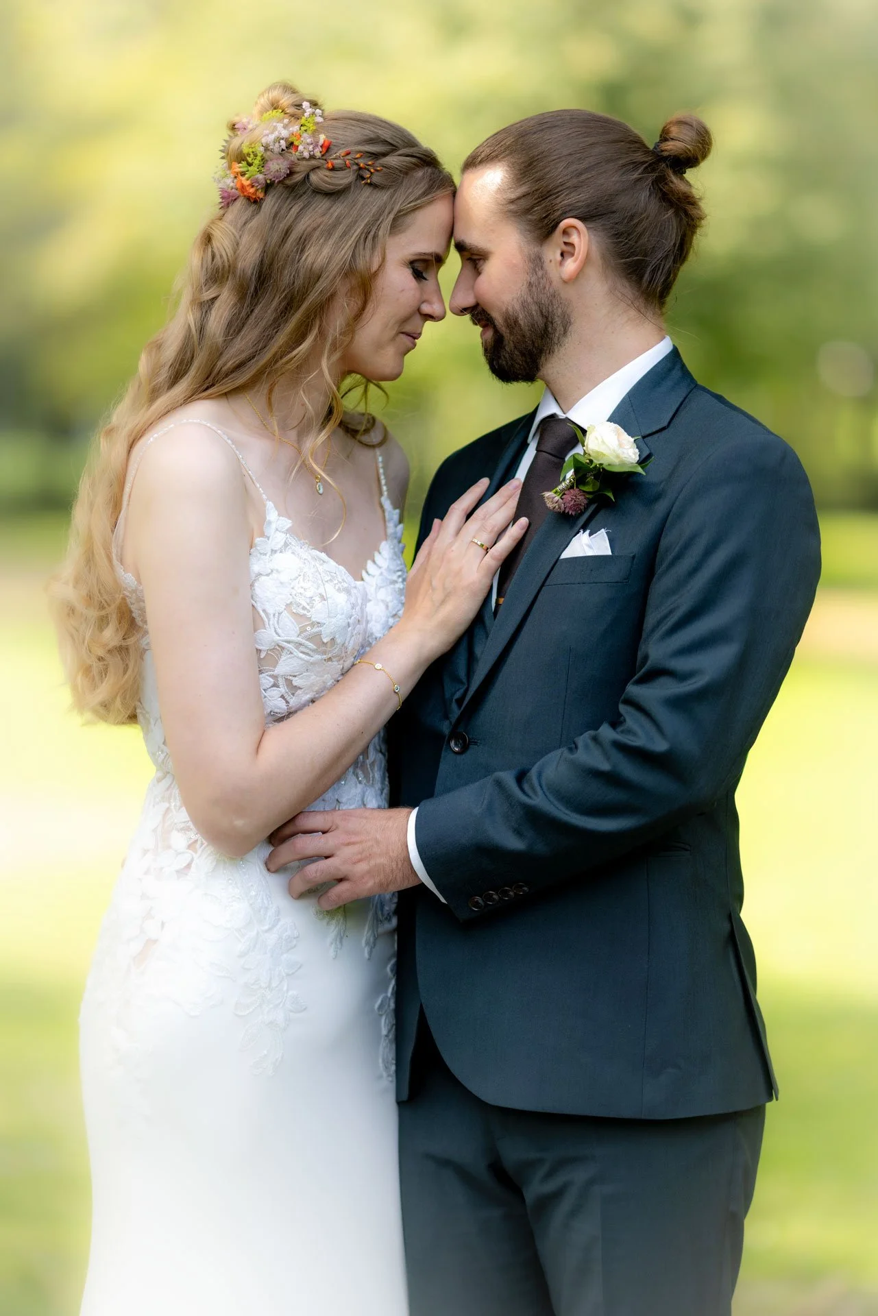 Et brudepar står tæt sammen udendørs, med panden mod hinanden, omgivet af grøn natur.