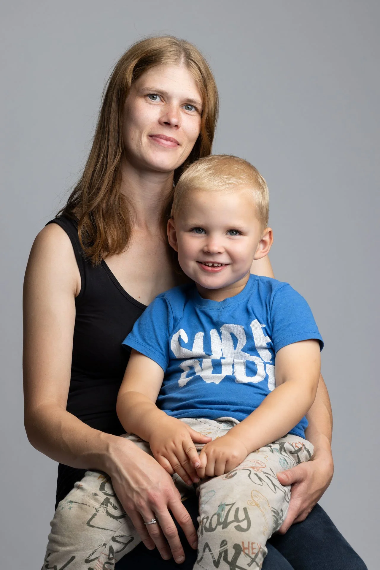 En kvinde holder en lille dreng på skødet, begge smiler mod kameraet, står foran en lysegrå baggrund.