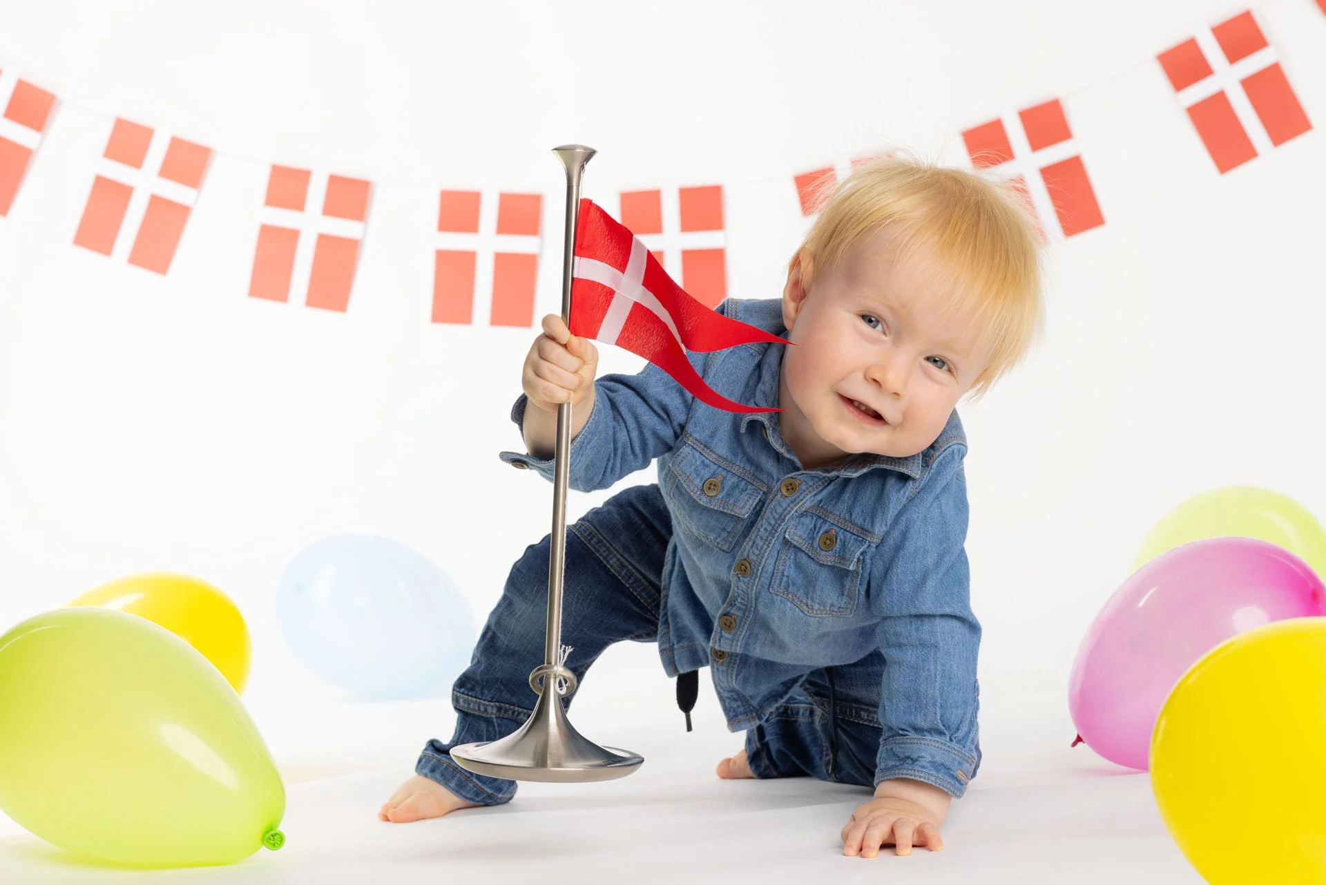 En glad ung dreng med blond hår iført denimjakke og jeans, der kryber på gulvet med en lille dansk flag på en stander, omgivet af farverige balloner og en dansk flag-bannercirkel i baggrunden.