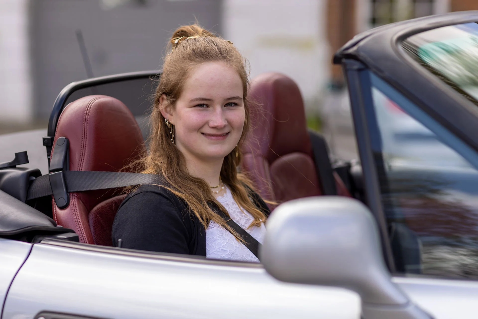 Ung kvinde med langt, brunt hår siddende i en cabriolet bil, smilende, med lilla og guld øreringe og en hvid bluse under en sort jakke.