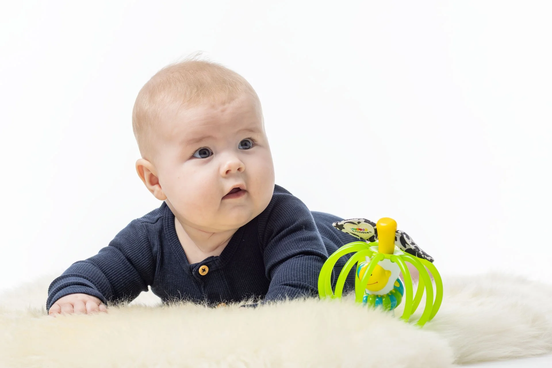 En baby ligger på maven på en blød, lys væg, med en legetøjsrangle foran sig.