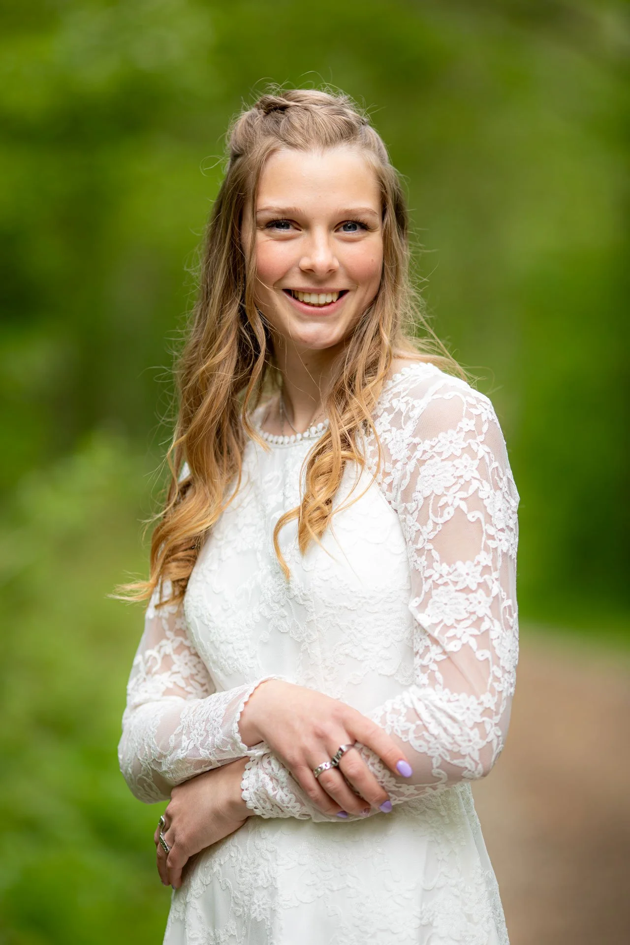 En smilende ung kvinde i en hvid blondekjole står i en grøn skov.