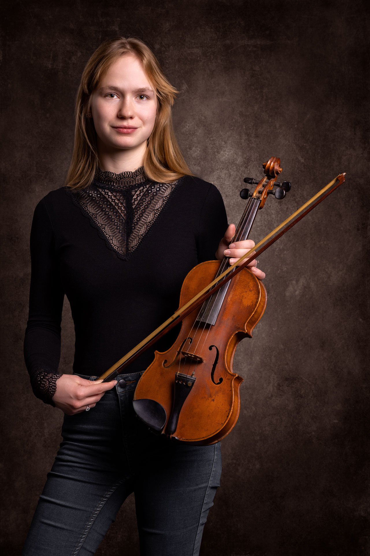 En kvinde med blond hår holder en violin og en violinbuet i hænderne, står mod en mørk, tekstureret baggrund.