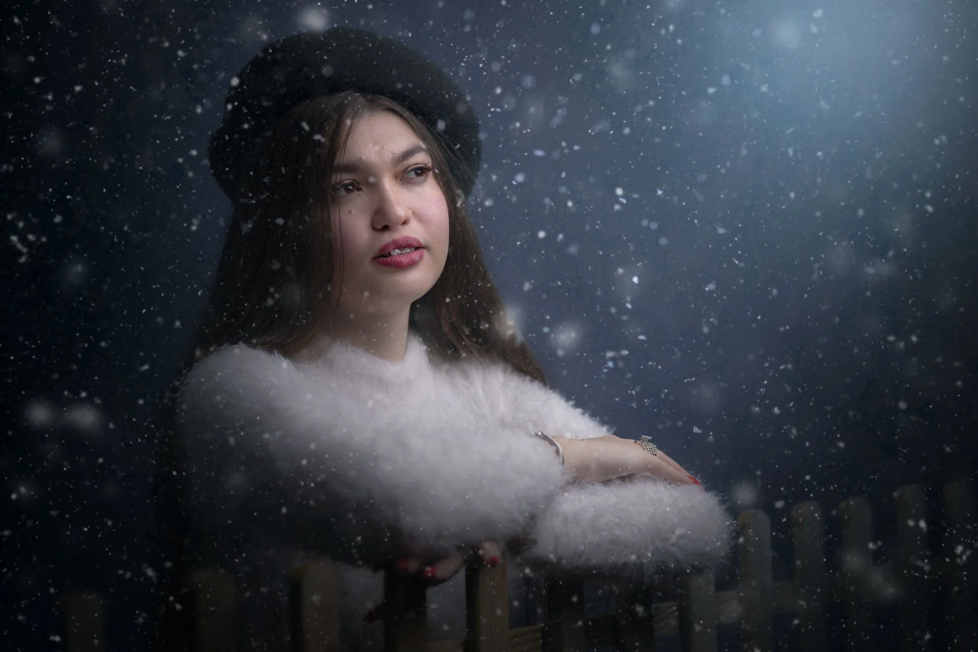En kvinde med langt brunt hår, iført en sort hat og en fluffy hvid sweater, står udenfor i sneen mod en mørk baggrund, med snefald omkring hende.