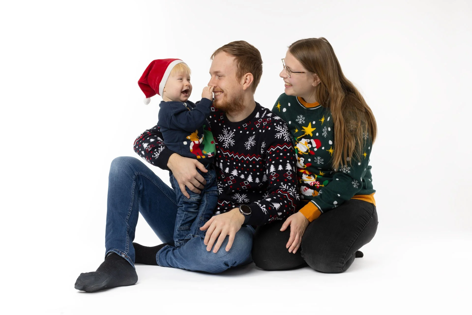 En familie med en far, mor og barn, alle iført juleleggings og juletrøjer, sidder på gulvet mod en hvid baggrund. Børn har julehætter. Familien smiler og leger med barnet.
