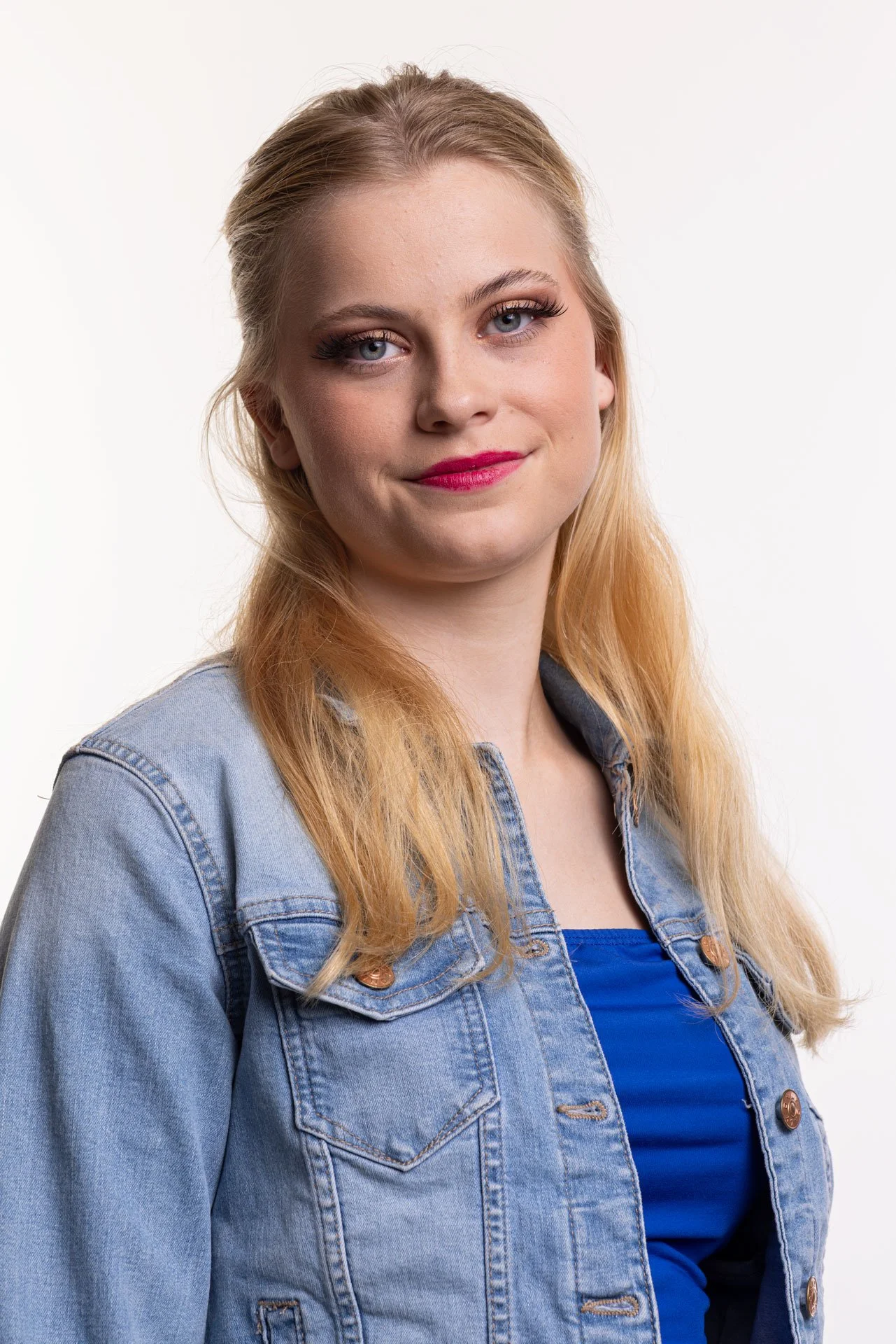 Kvindelig portræt med blondt hår, blå øjne, smilende, iført denimjakke og blå top, mod hvid baggrund.