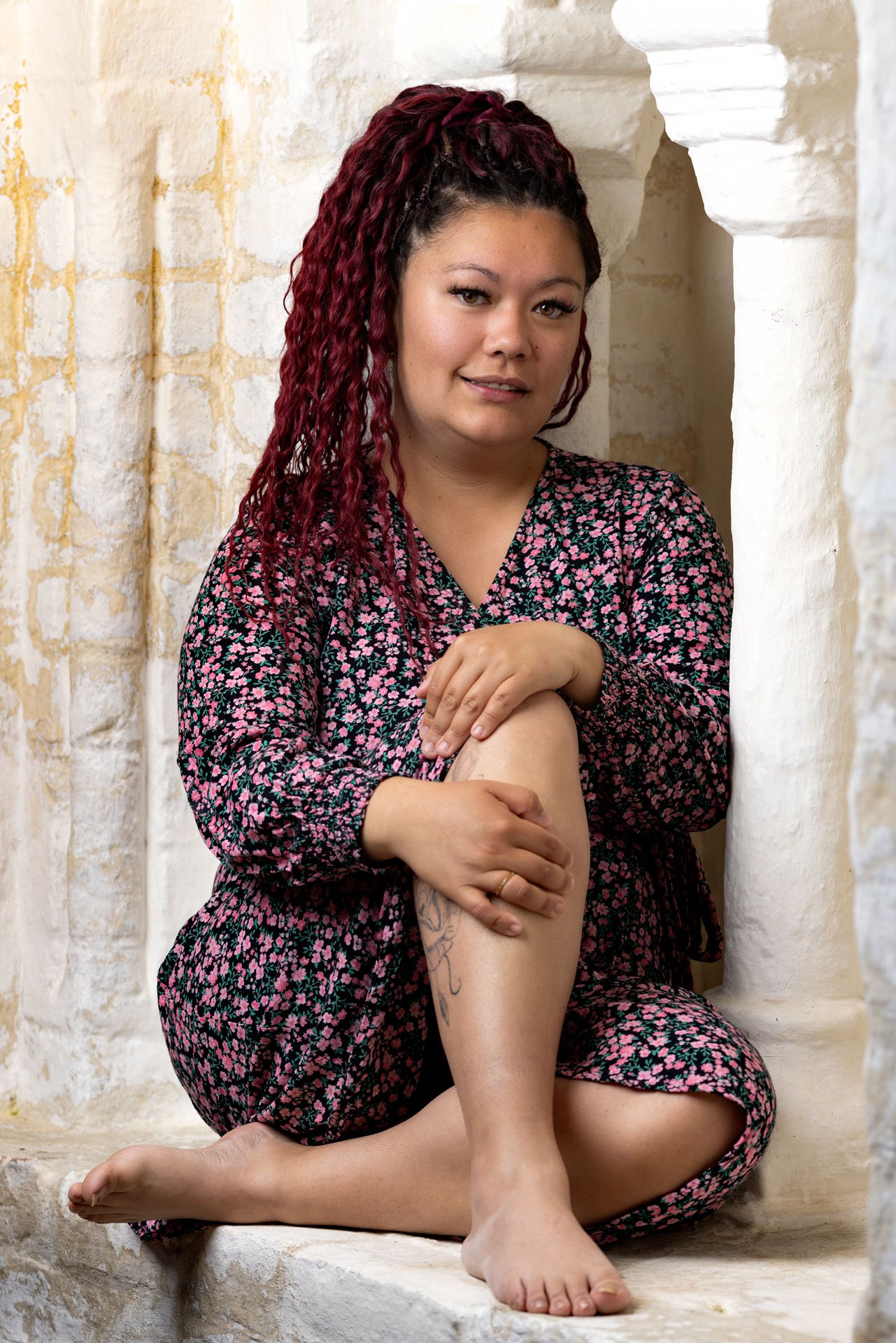 Kvinde med langt, krøllet, mørkrødt hår sidder på knæ i en murstensbue, iført en sort kjole med pink blomster, og viser sin tatovering på benet.