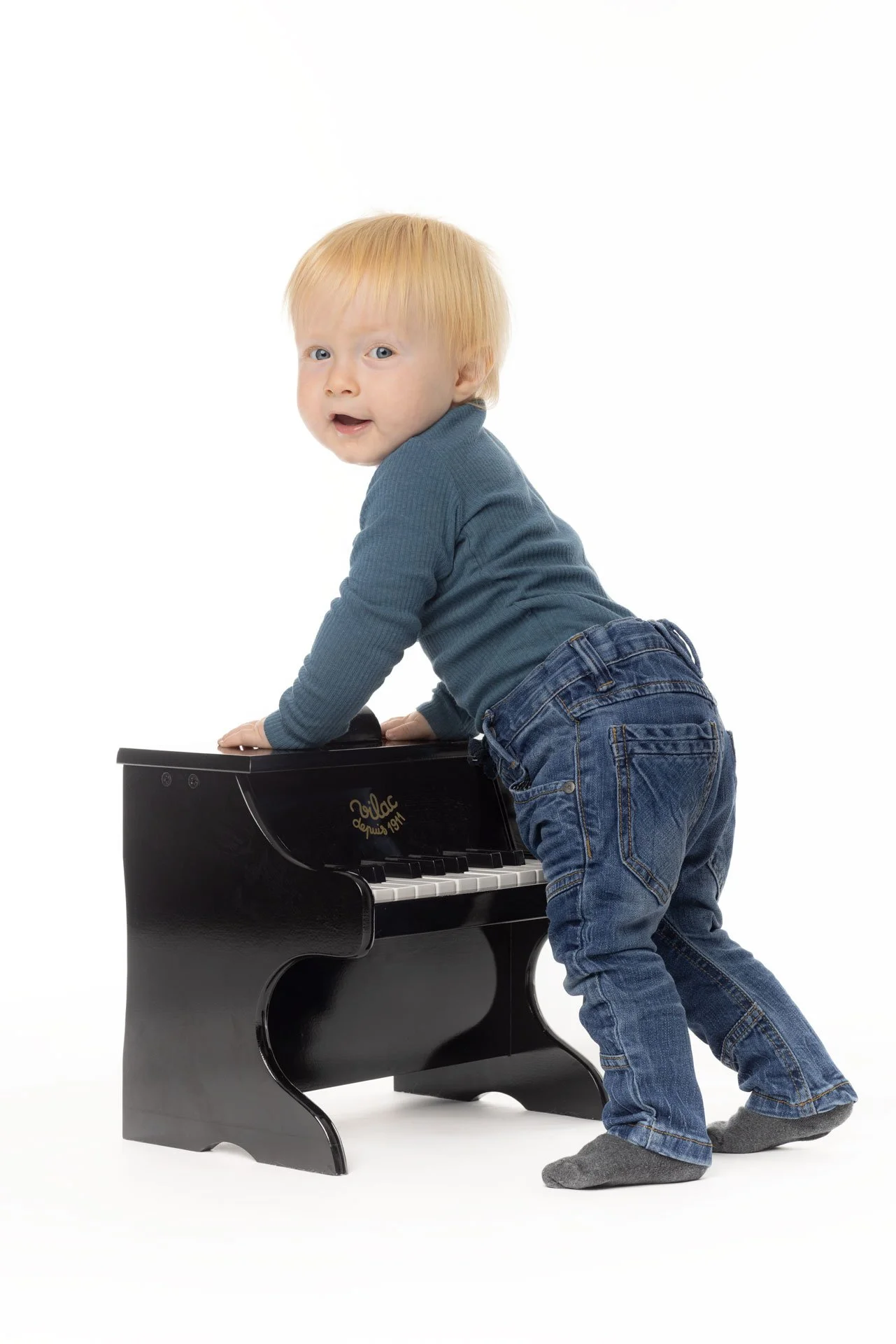 En ung dreng med blond hår, iført en blå langærmet trøje og jeans, står på alle fire og leger med en lille sort klaver mod en hvid baggrund.