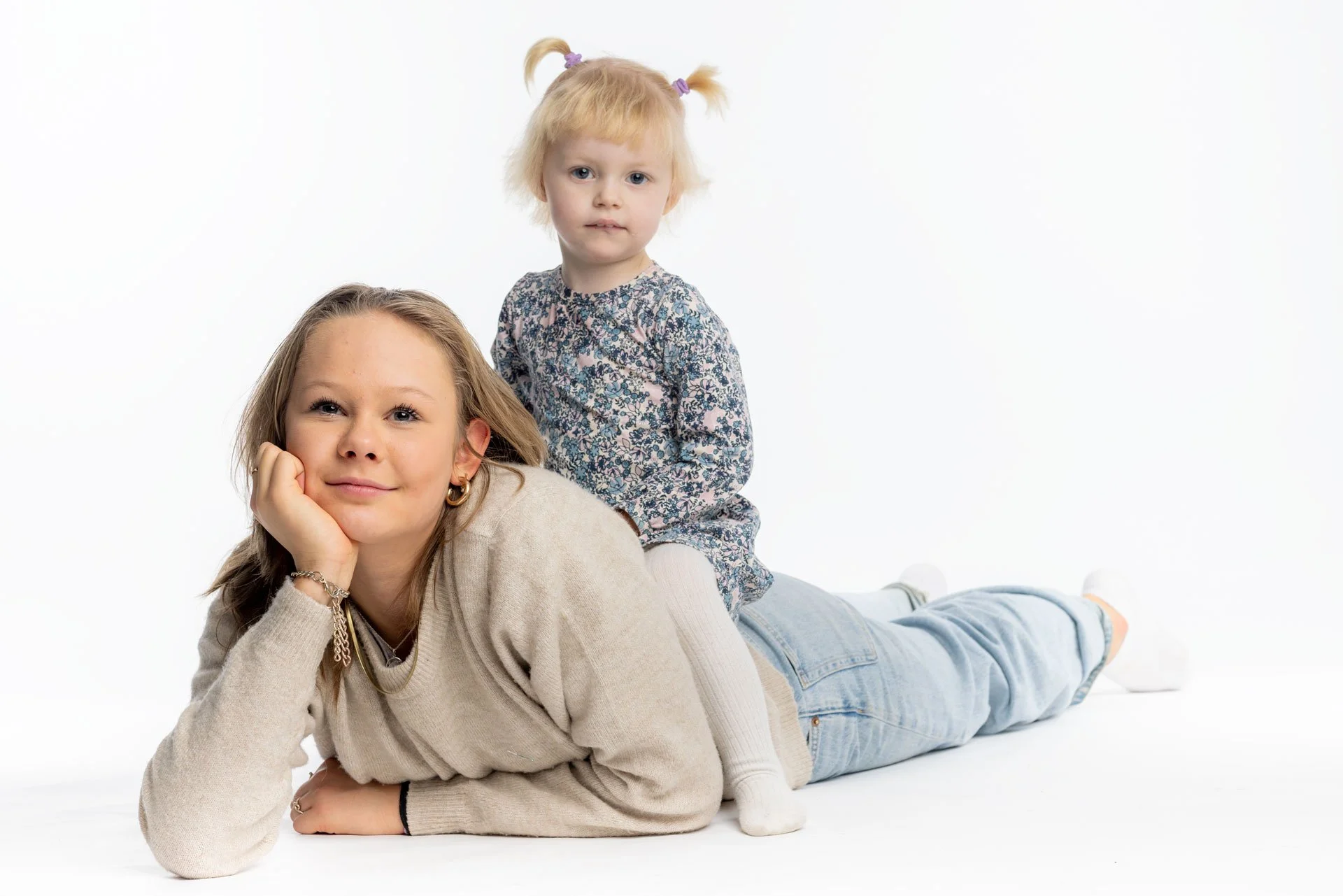 En voksen kvinde ligger ned med hovedet hvilede i hendes hånd, mens en lille pige sidder på hendes ryg. Begge har lyst hår og smilende ansigter, og baggrunden er hvid.