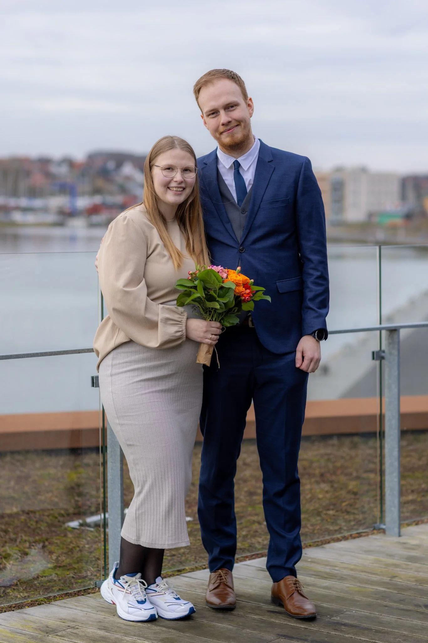 En kvinde og en mand står sammen på en udendørs café ved vandet, kvinden holder en blomsterbuket, begge smiler og ser mod kameraet, med bybebyggelse i baggrunden.