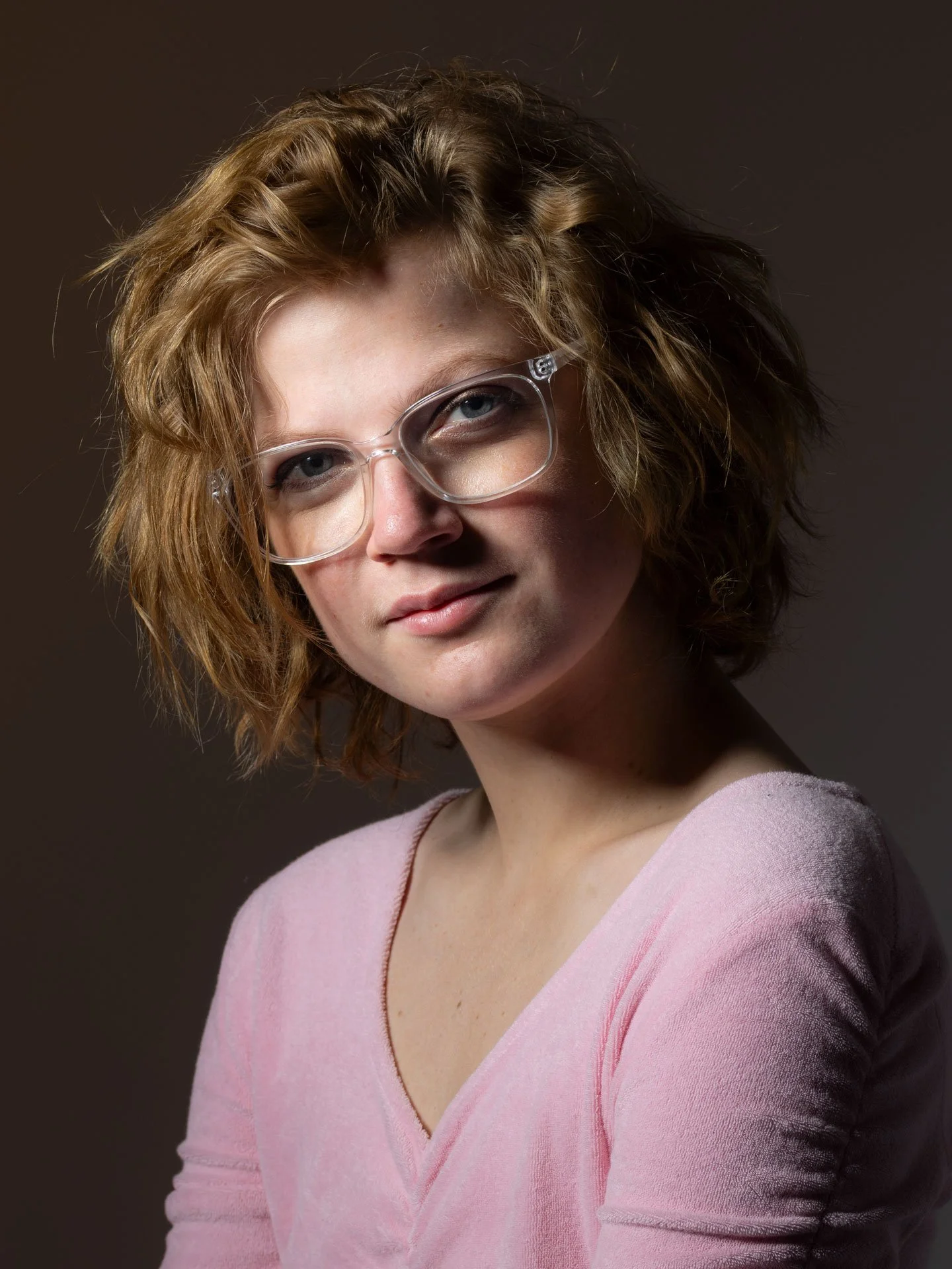 Portræt af en kvinde med lyst, krøllet hår iført klare briller og en lyserød top, der smiler let til kameraet.