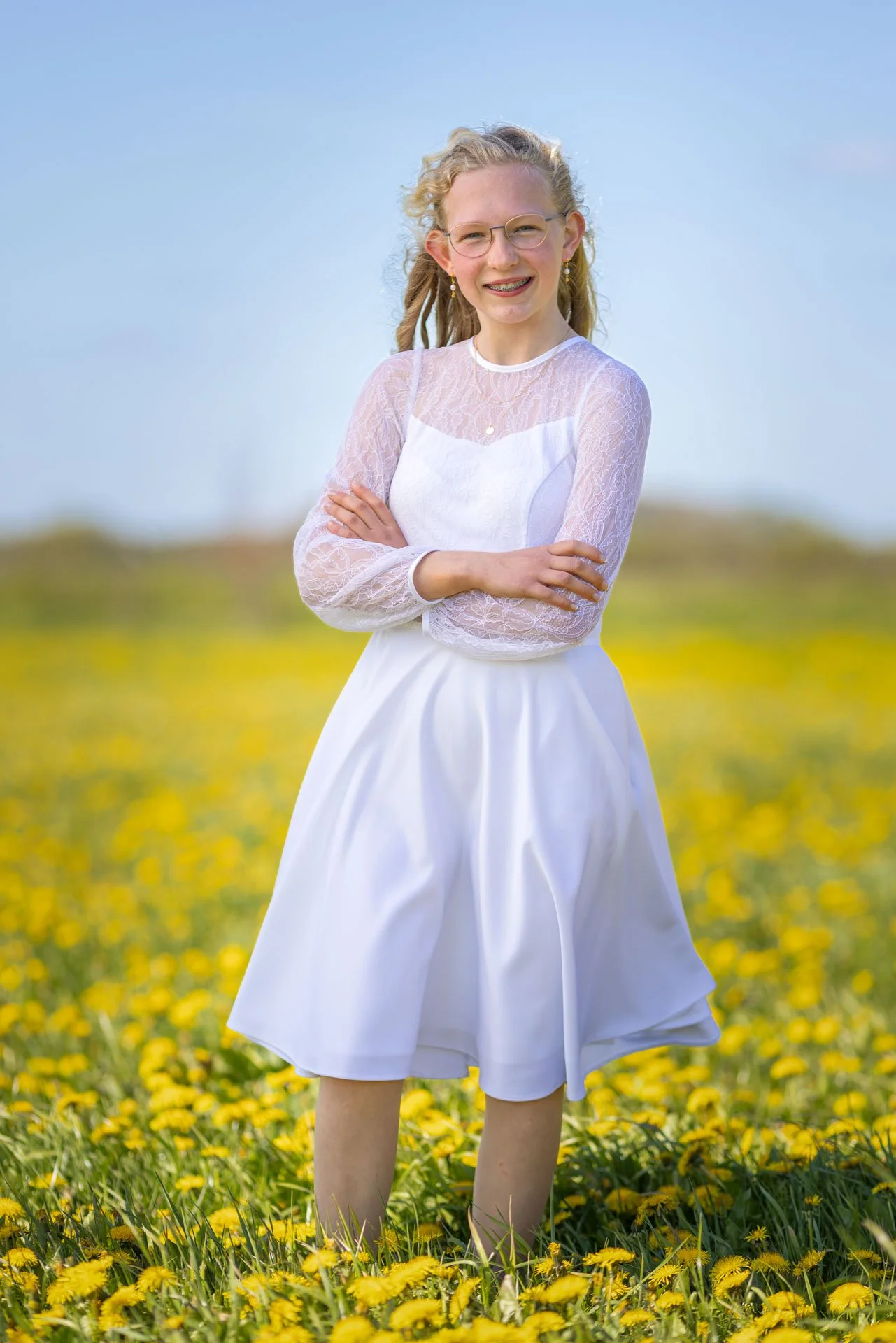 Smilende ung pige i hvid kjole står i en mark med gule blomster under en blå himmel.