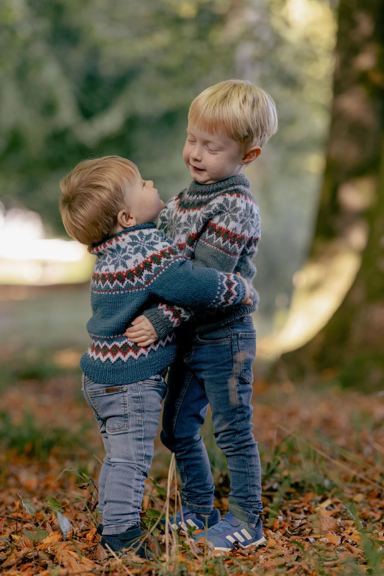To unge drenge i jakkesæt, der leger i en skov, iført varme strikkede sweater med julemotiver.