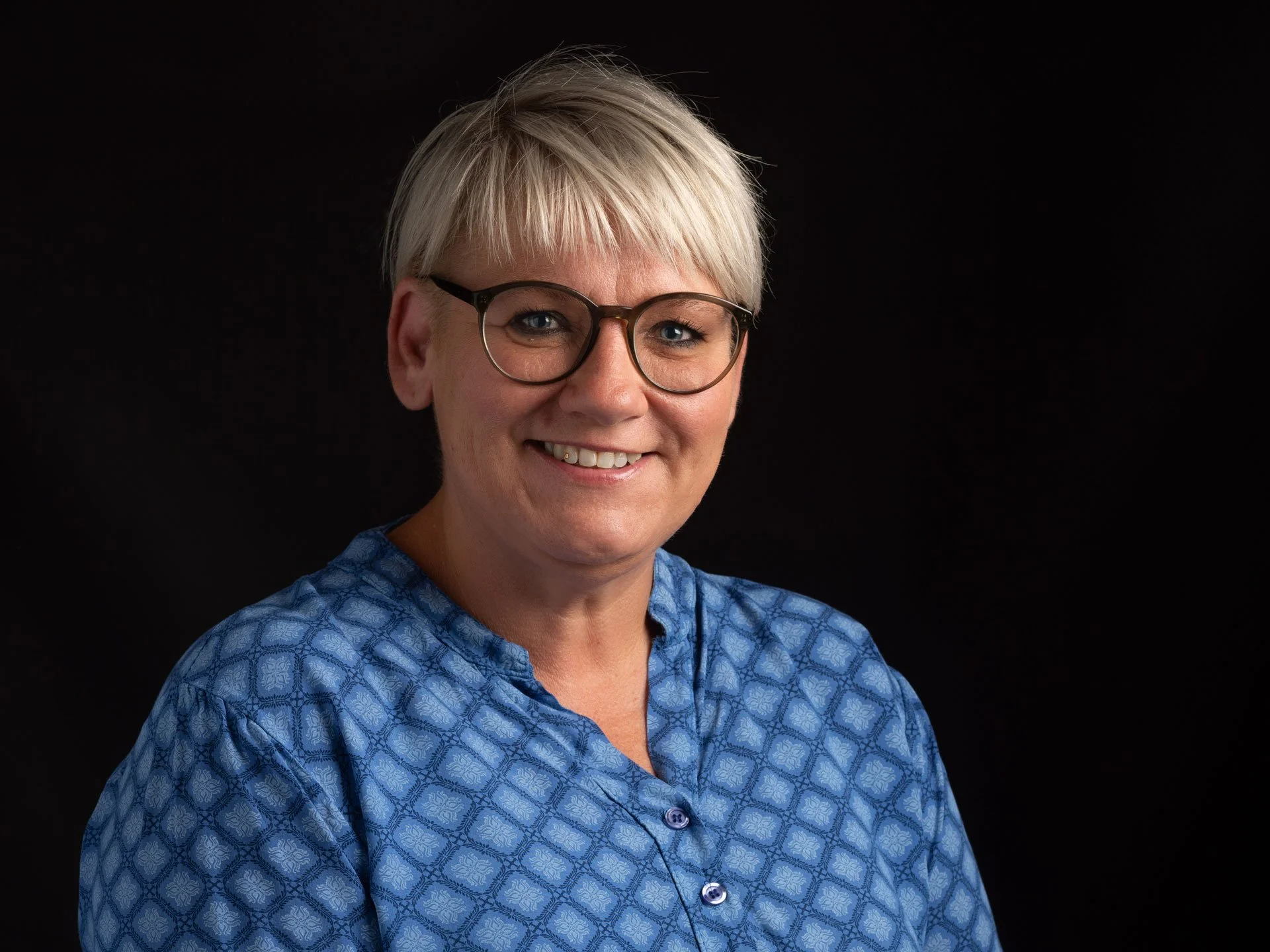 Et portræt af en smilende kvinde med kort blond hår, briller, og en blå bluse med et mønster, mod en sort baggrund.