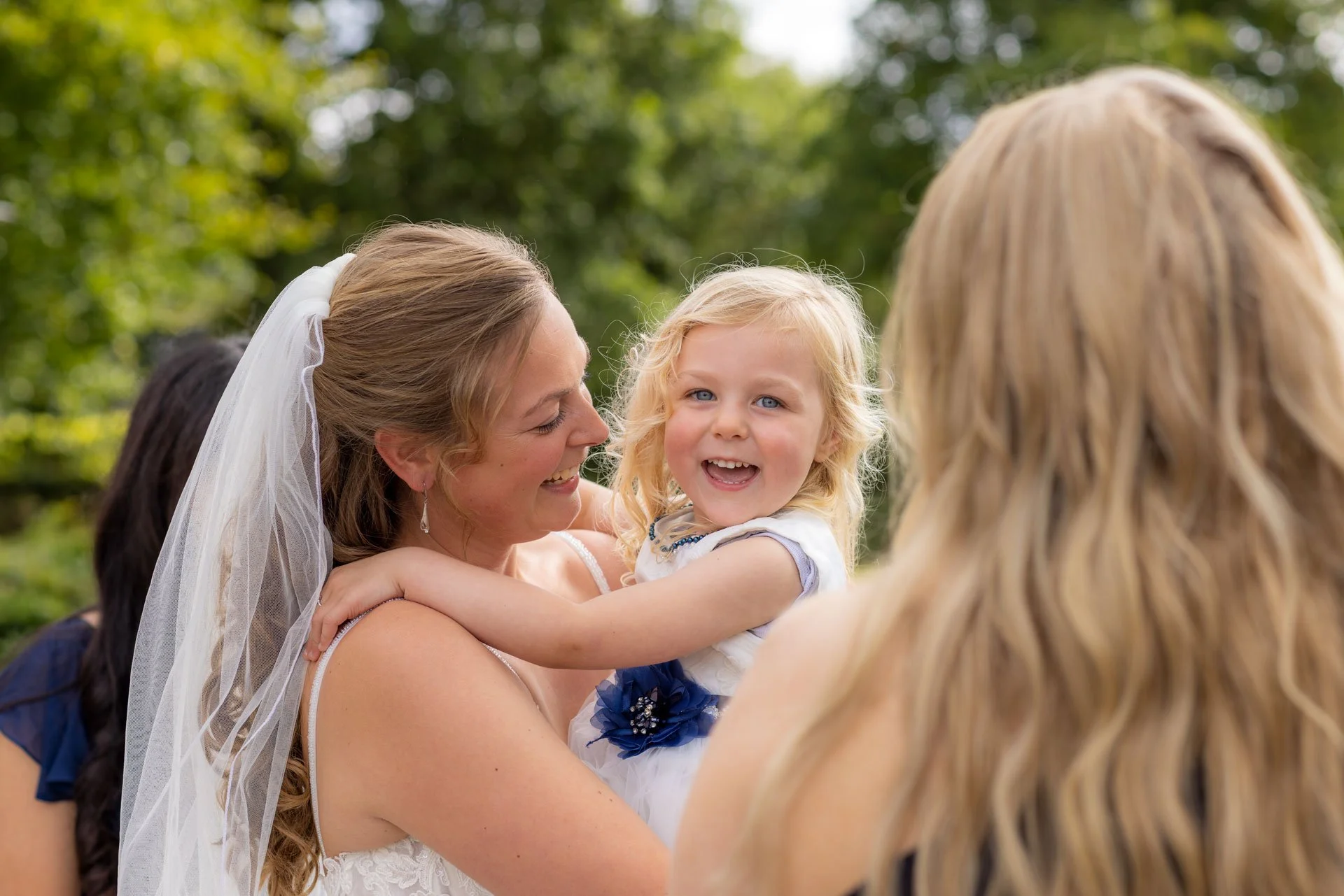 En kvinde med brunt hår og en brudekjole holder en lille pige med blond hår og blå øjne, som smiler og har armene om hendes hals. De er udenfor med grønne træer i baggrunden.