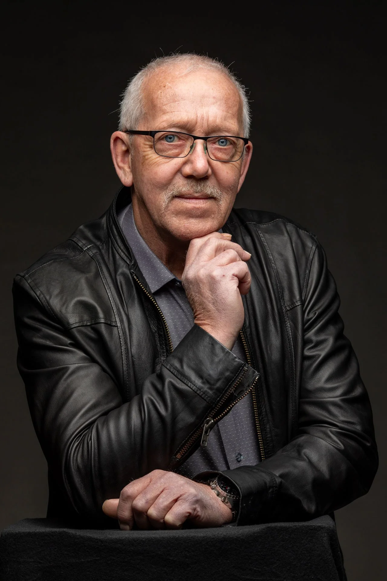 Et portræt af en ældre mand med brunt, tyndt hår, iført briller, sort læderjakke og en grå skjorte, der sidder med hånden under hagen mod en sort baggrund.