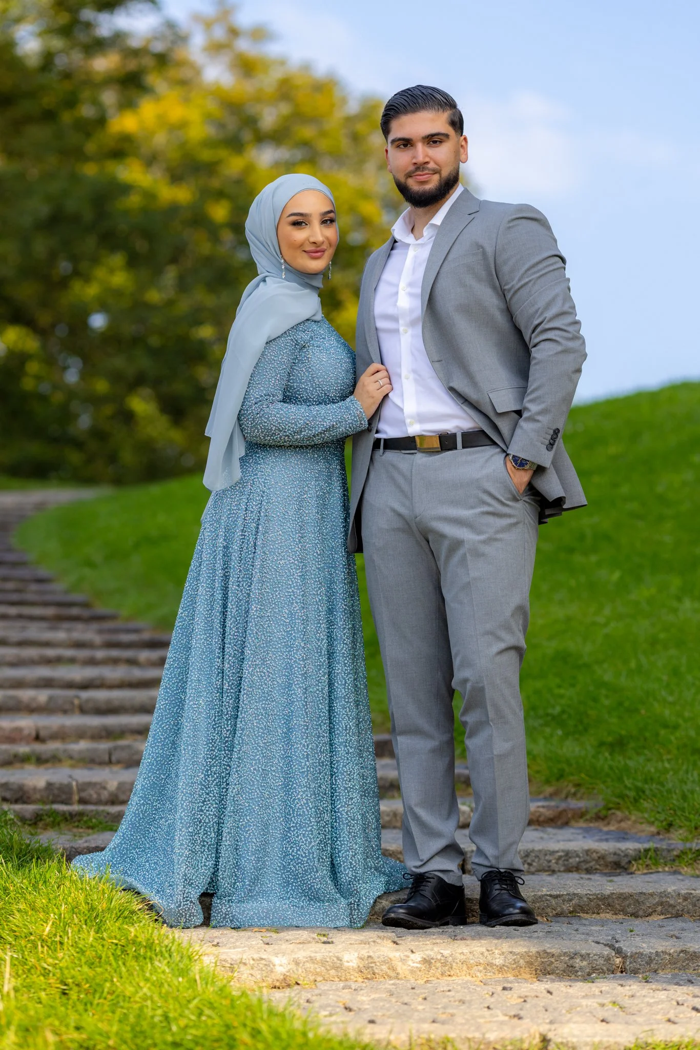 Et par står på en sti foran grønne buske og træer, med en blå himmel i baggrunden, klædt i formelt tøj, kvinden i en lang, blåt kjole med tørklæde, og manden i en grå jakkesæt.