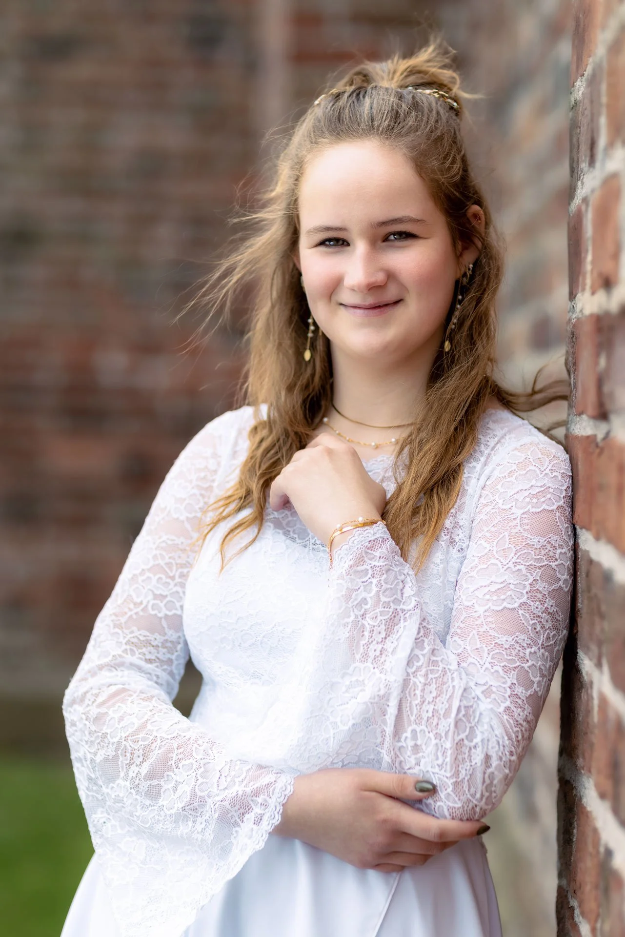 Ung kvinde med lang, let krøllet hår staar ved en mur af sten, iført en hvid kjole med blondebånd og smykker, smilende mod kameraet.