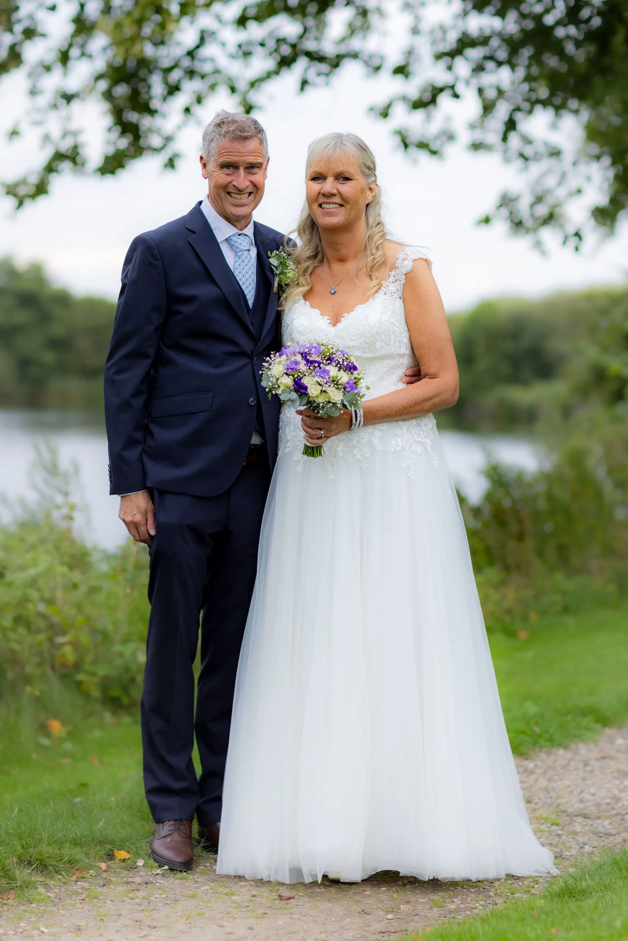 Et brudepar står udendørs ved en sø, iført bryllupsklar tøj, smilende til kameraet, med grønne træer og en sø i baggrunden.