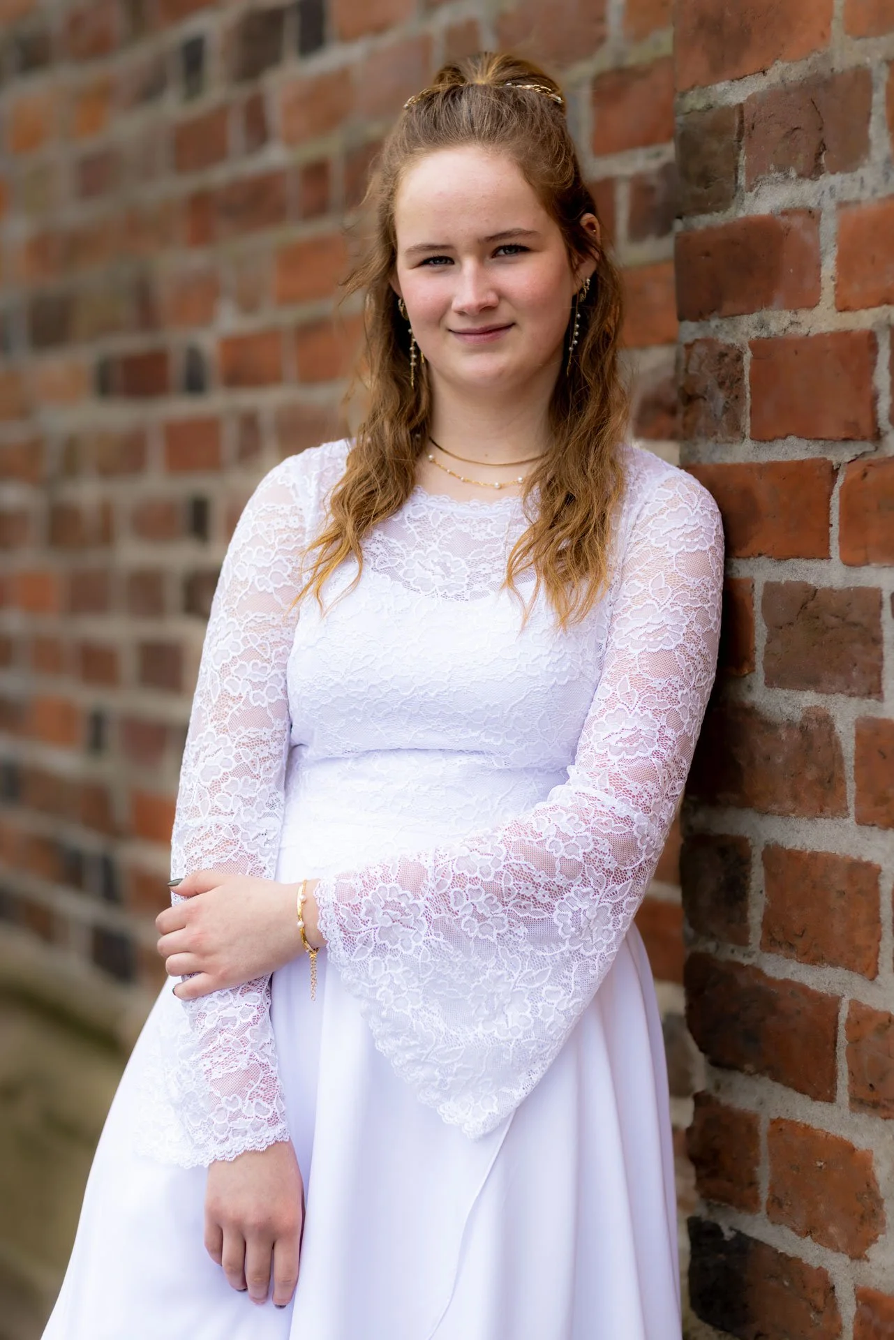 En ung kvinde med brunt, bølget hår, iført en hvid brudekjole med blondeærmer, står op ad en murstensmur.