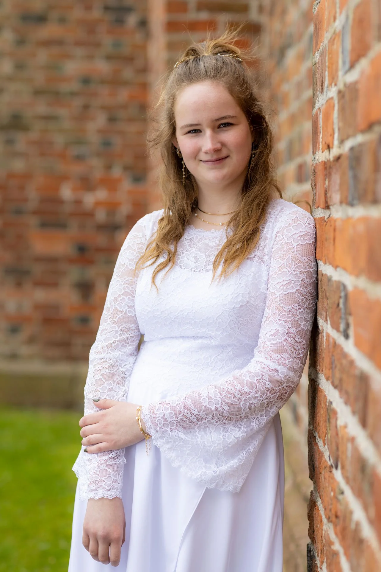 En ung kvinde med langt, krøllet, blond hår står ved en mur, iført en hvid, blondelignende kjole. Hun smiler let og har smykker på, herunder øreringe, halskæde og armbånd.