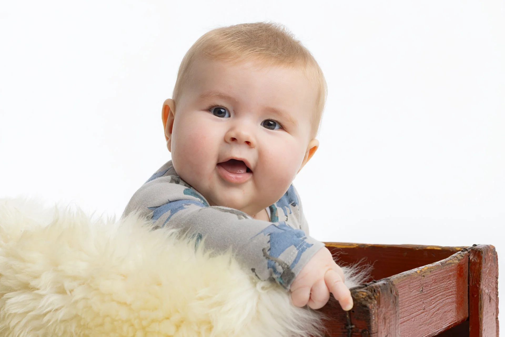 Sød baby med lyst hår, der leger på en blød cremefarvet pels, med en gammel trækasse mod en hvid baggrund.