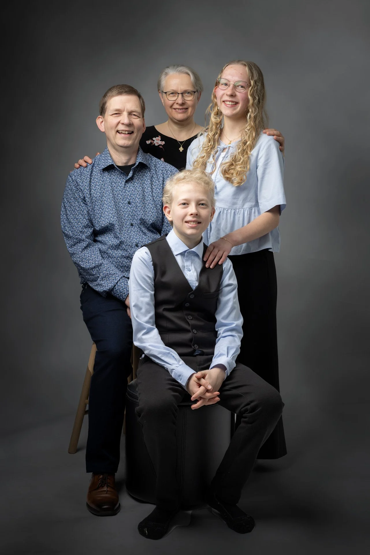 En familie portræt med fire personer mod en grå baggrund. En kvinde står bag hendes mand, mens en teenage pige står til højre. En dreng sidder foran på en sort sæde stol.