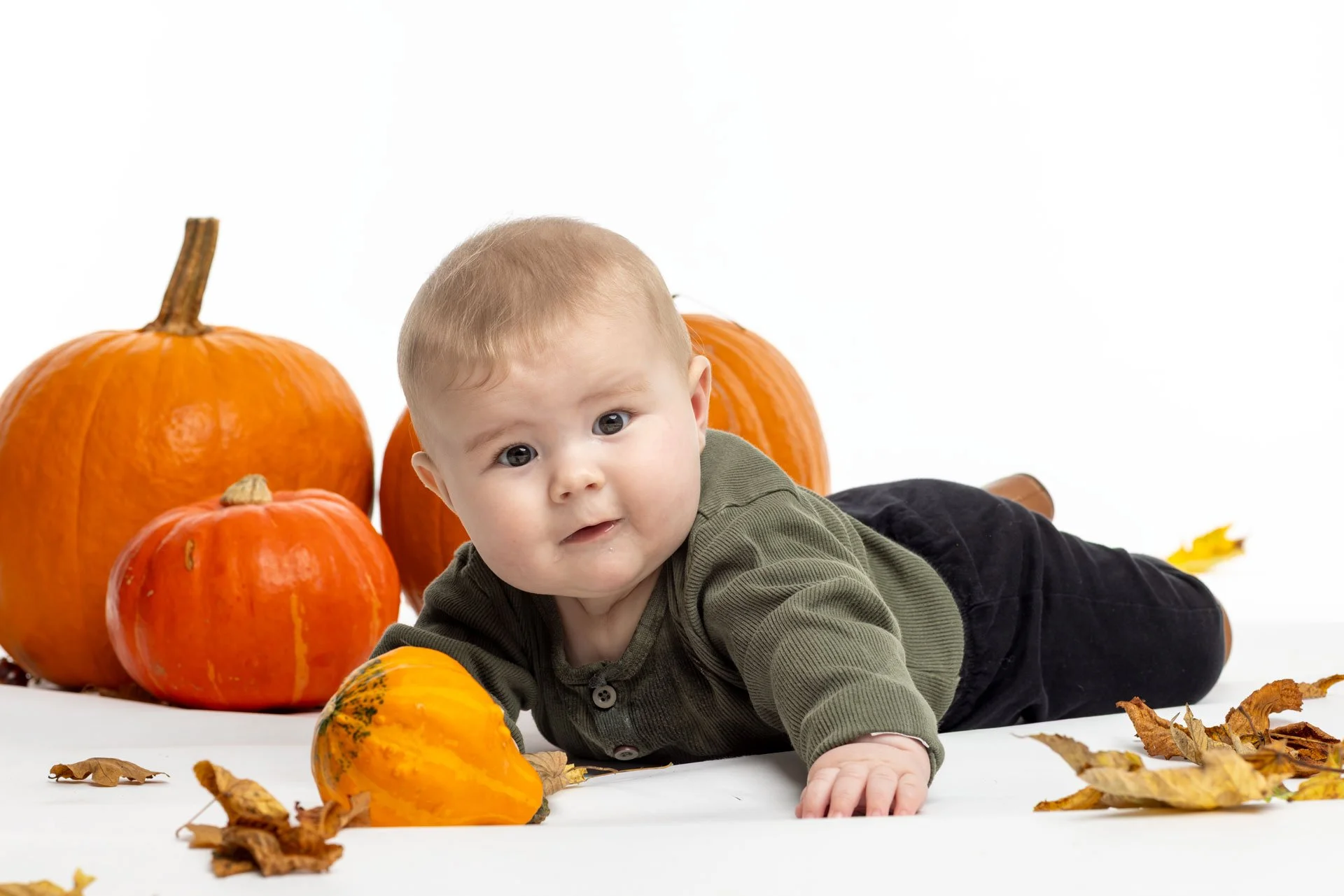 En baby ligger på maven omgivet af efterårsgrene og flere orange græskar.