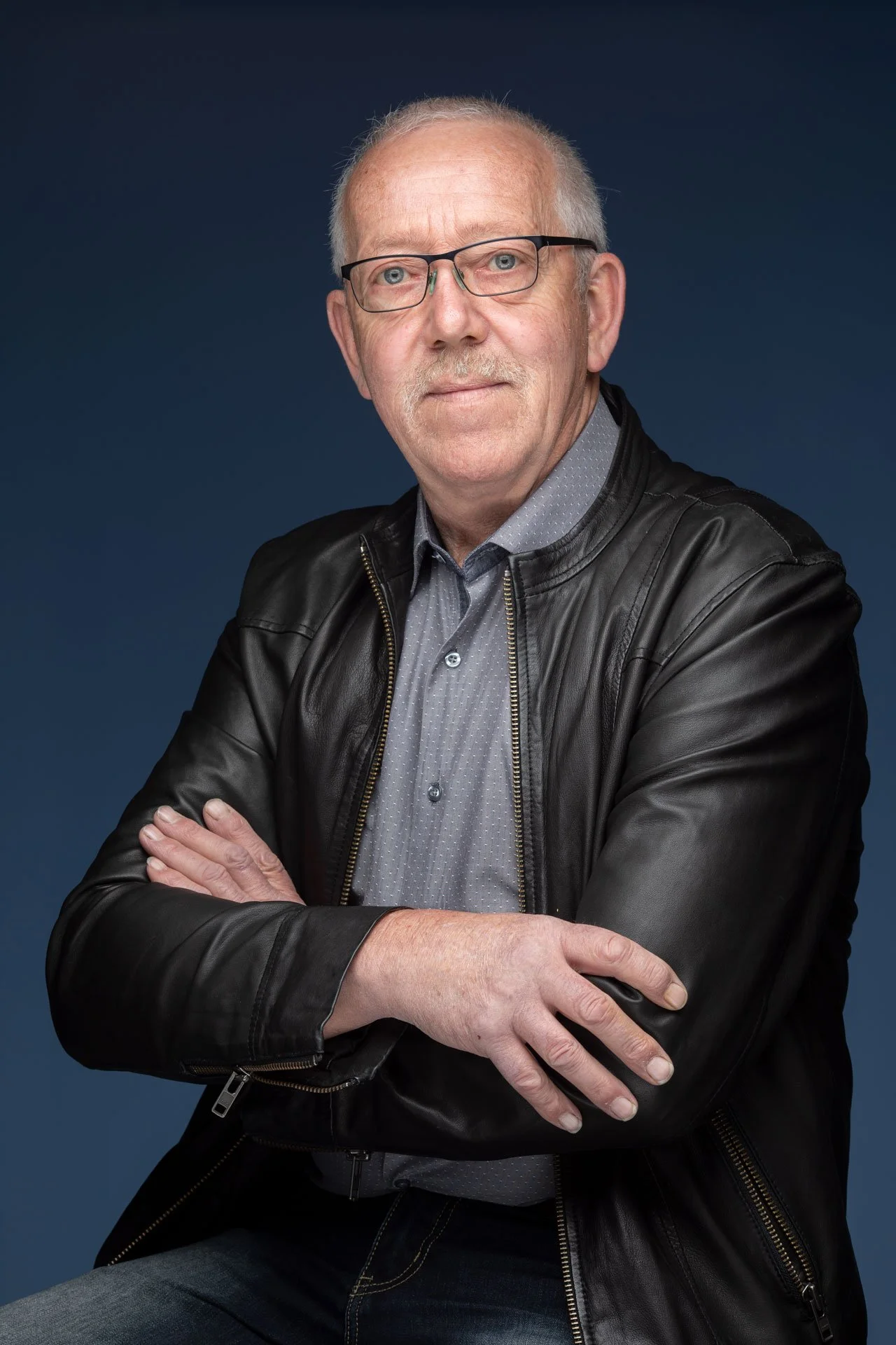 Portræt af ældre mand med briller, iført sort læderjakke og lyse skjorte mod en mørk blå baggrund.