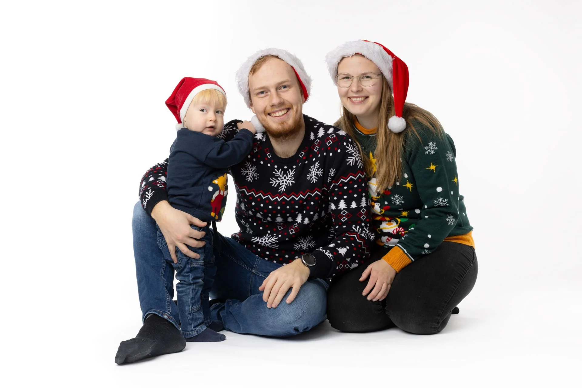 Familie på tre iført juletrøjer og nissehuer, smilende, på en hvid baggrund.