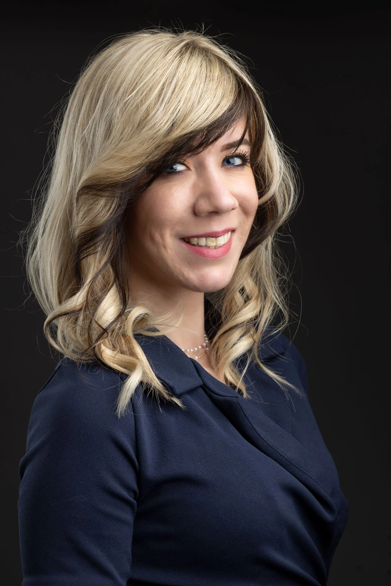 Portræt af en smilende kvinde med blond, bølget hår, iført en mørk jakke og perlekæde, mod en sort baggrund.