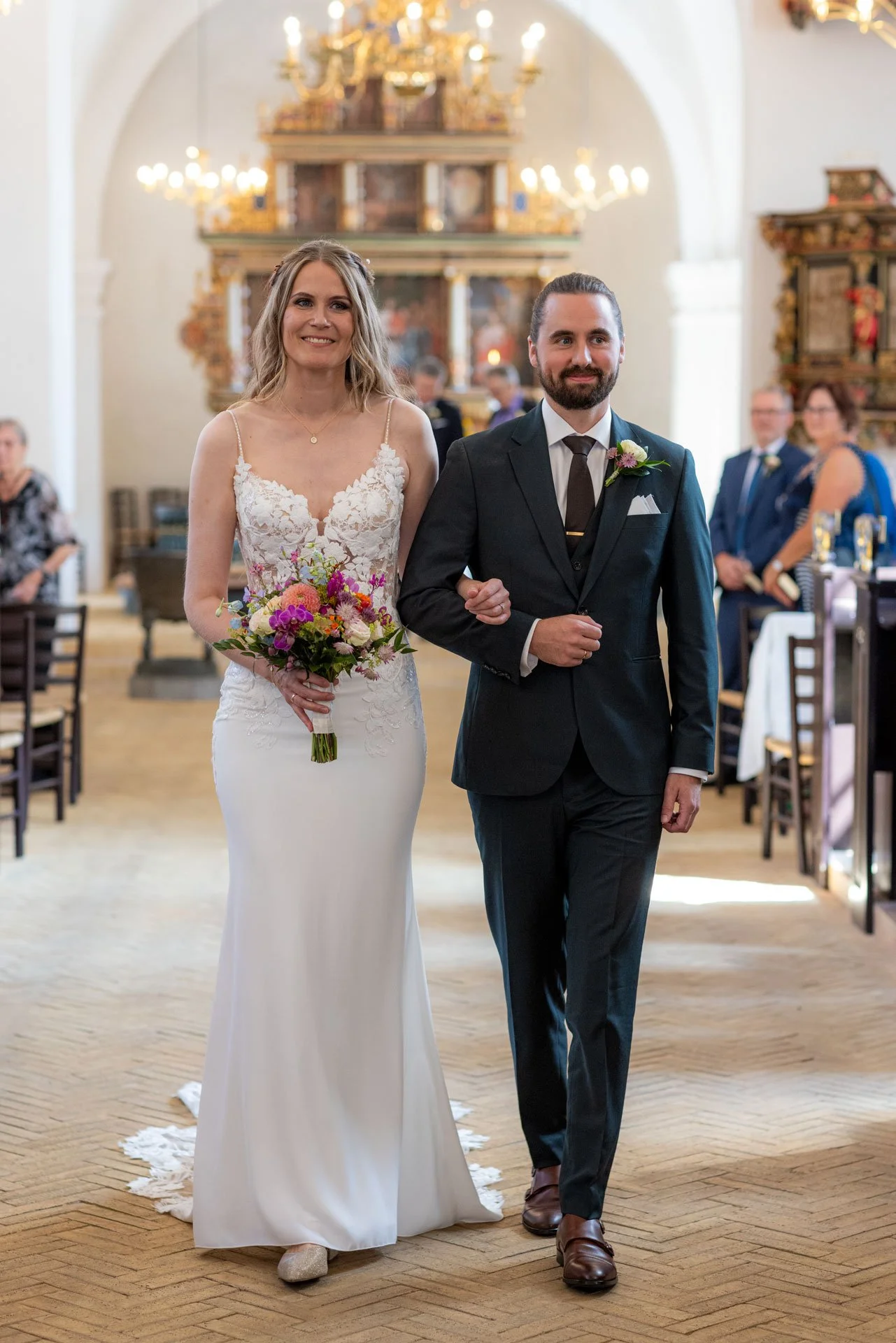 Nygifte par går ned ad kirkegulvet under et bryllup i en kirke, omgivet af gæster.